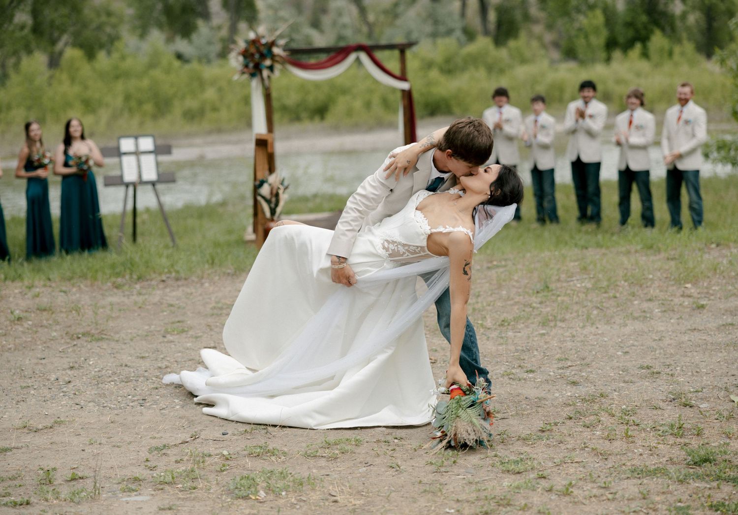 Groom dips bride in a romantic kiss at outdoor wedding ceremony with wedding party standing in background.