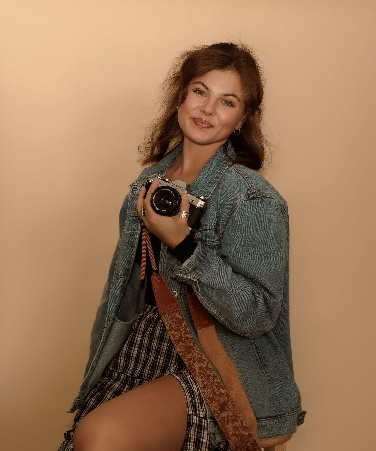 Smiling young woman in denim jacket and plaid skirt posing with a camera against a beige studio background.