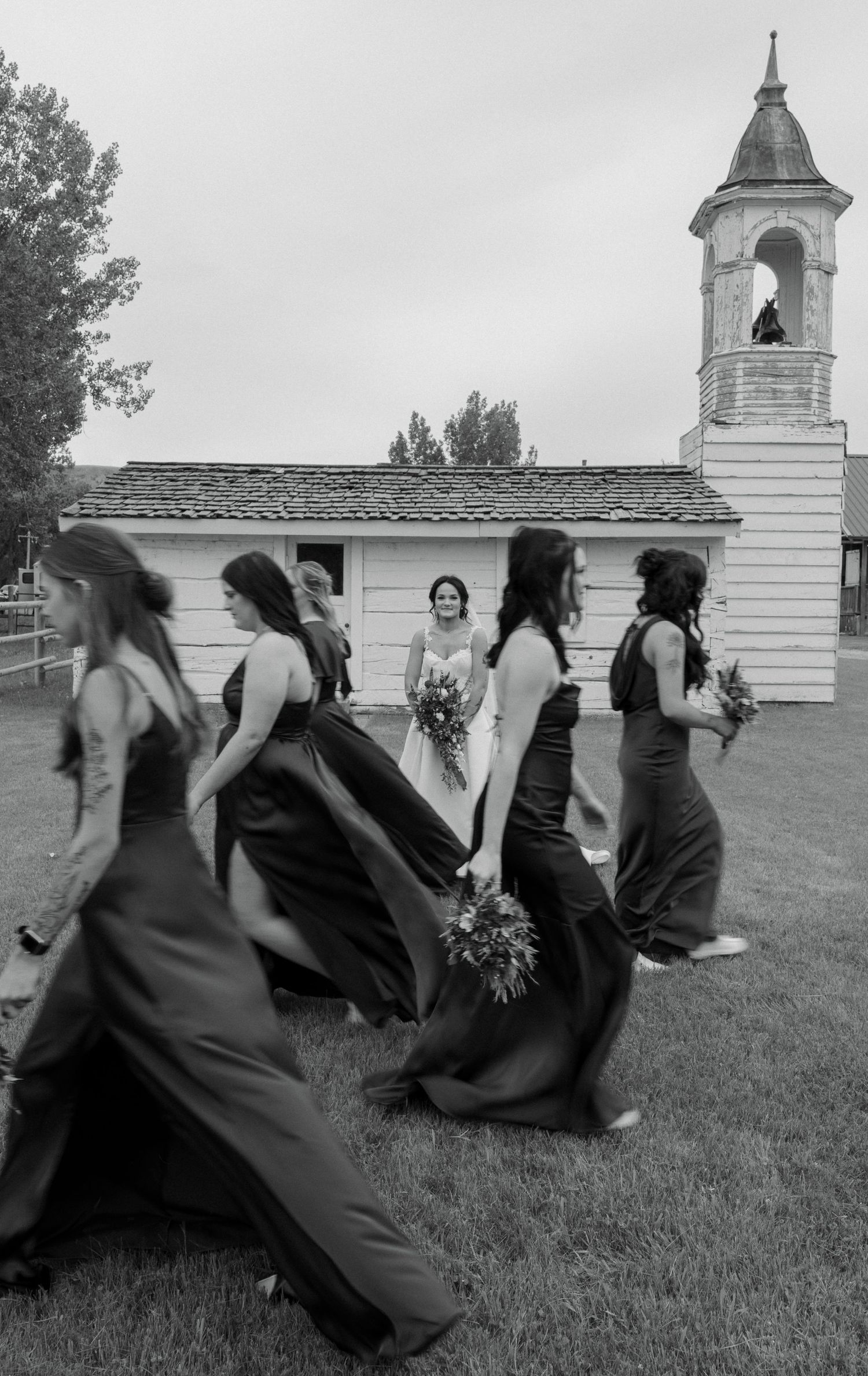 Black and white photo of bridesmaids in dark gowns walking past a small white chapel, bride visible in background holding bouquet.