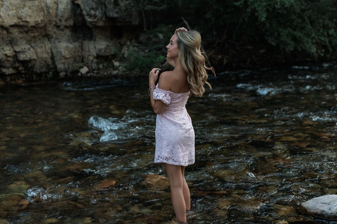 Young blonde woman in pink lace off-shoulder dress standing in rocky mountain stream.