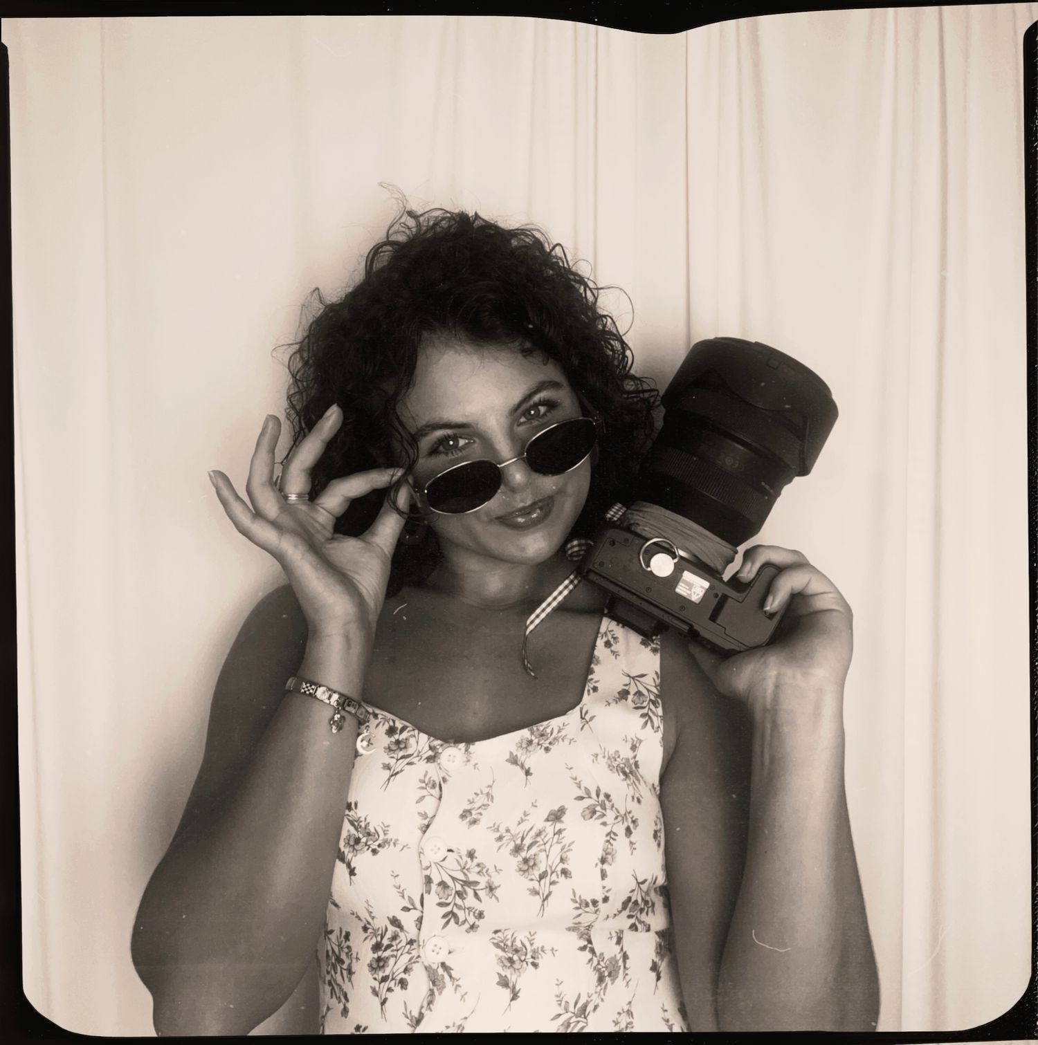 Black and white photo booth portrait of a curly-haired woman in sunglasses holding a vintage camera.