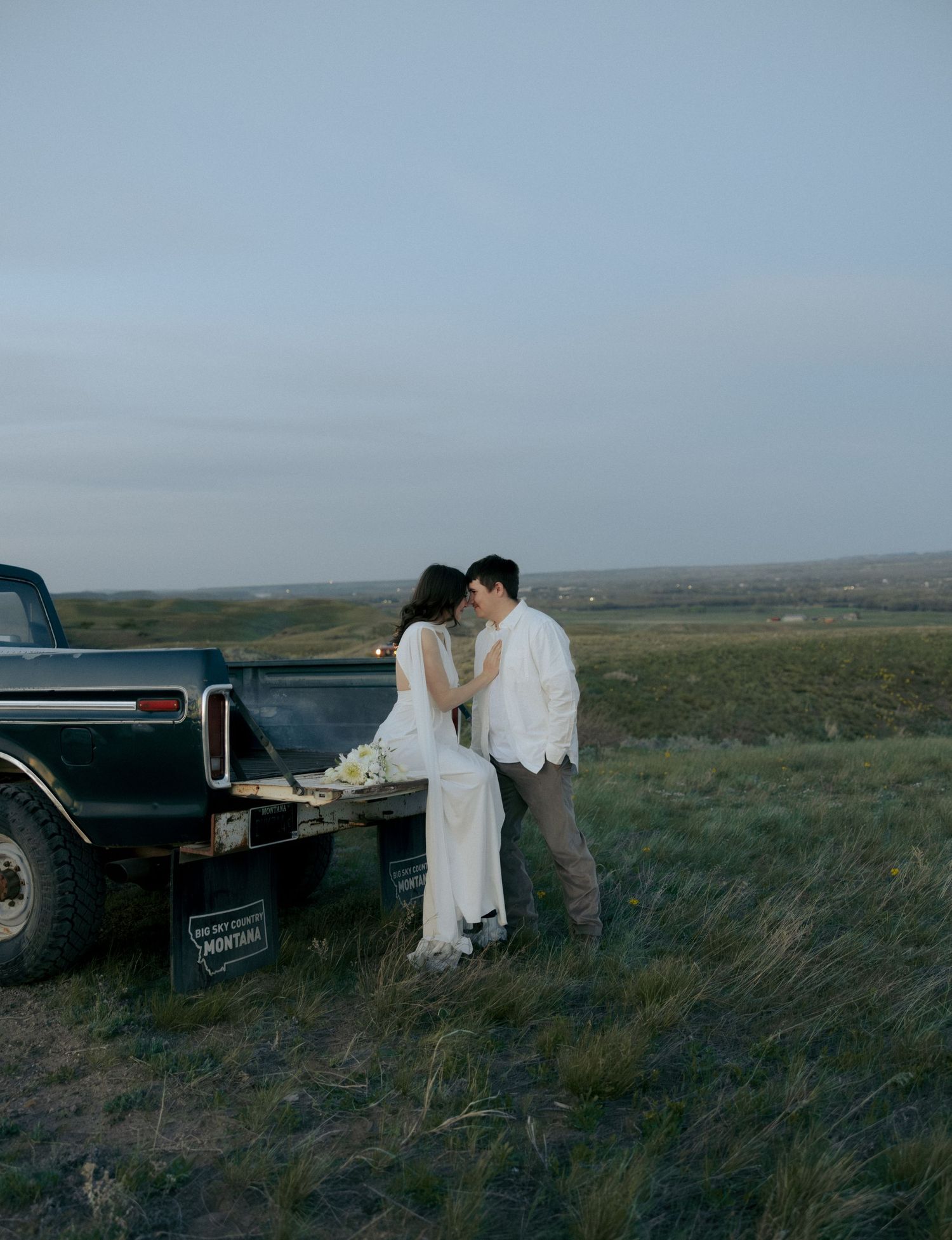 A couple in wedding attire shares a romantic moment leaning against a vintage truck in an open field at dusk.