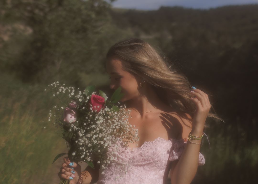 Young woman in pink off-shoulder dress smelling colorful flower bouquet outdoors.