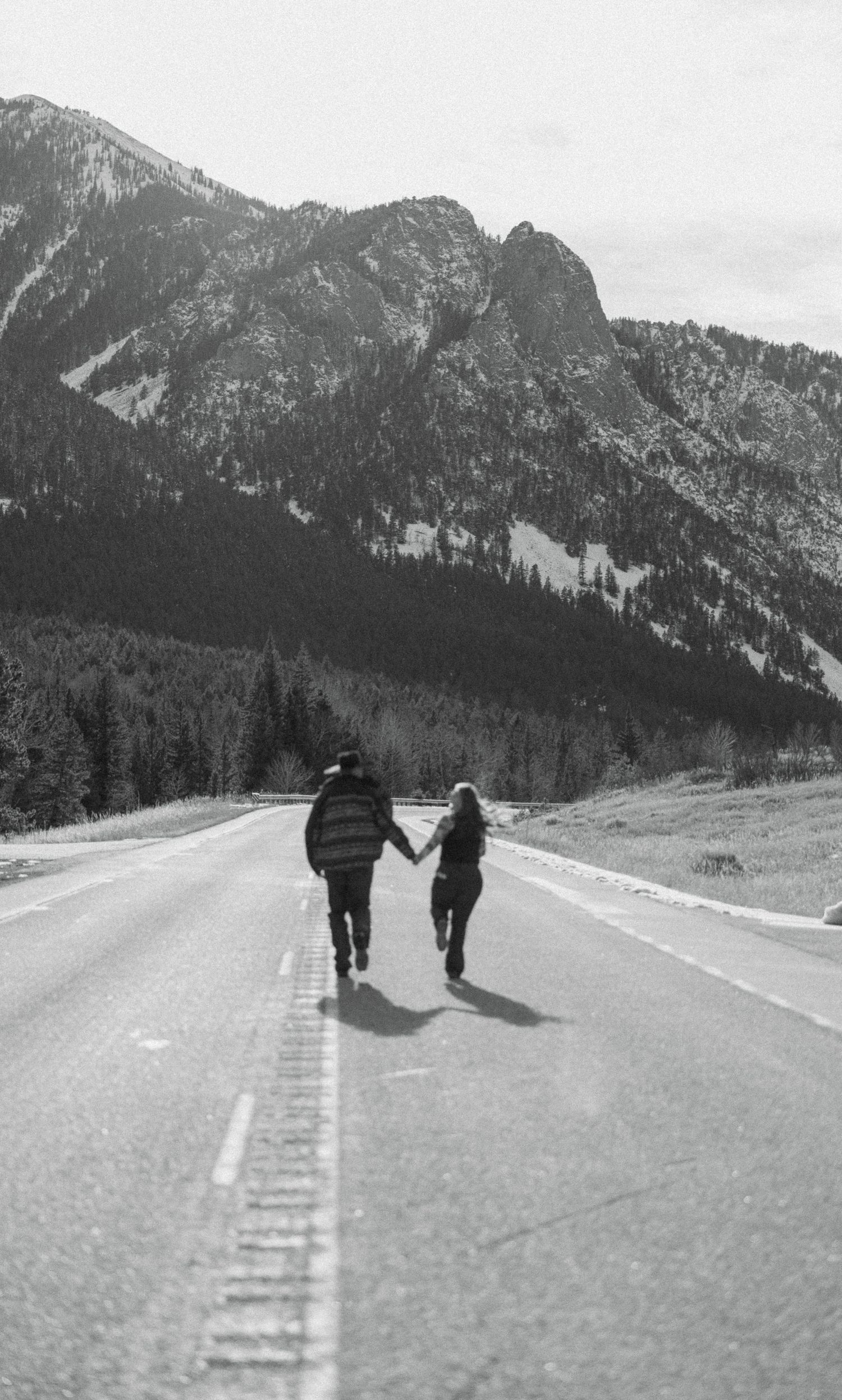 Black and white photo of couple holding hands running down an open mountain highway together.