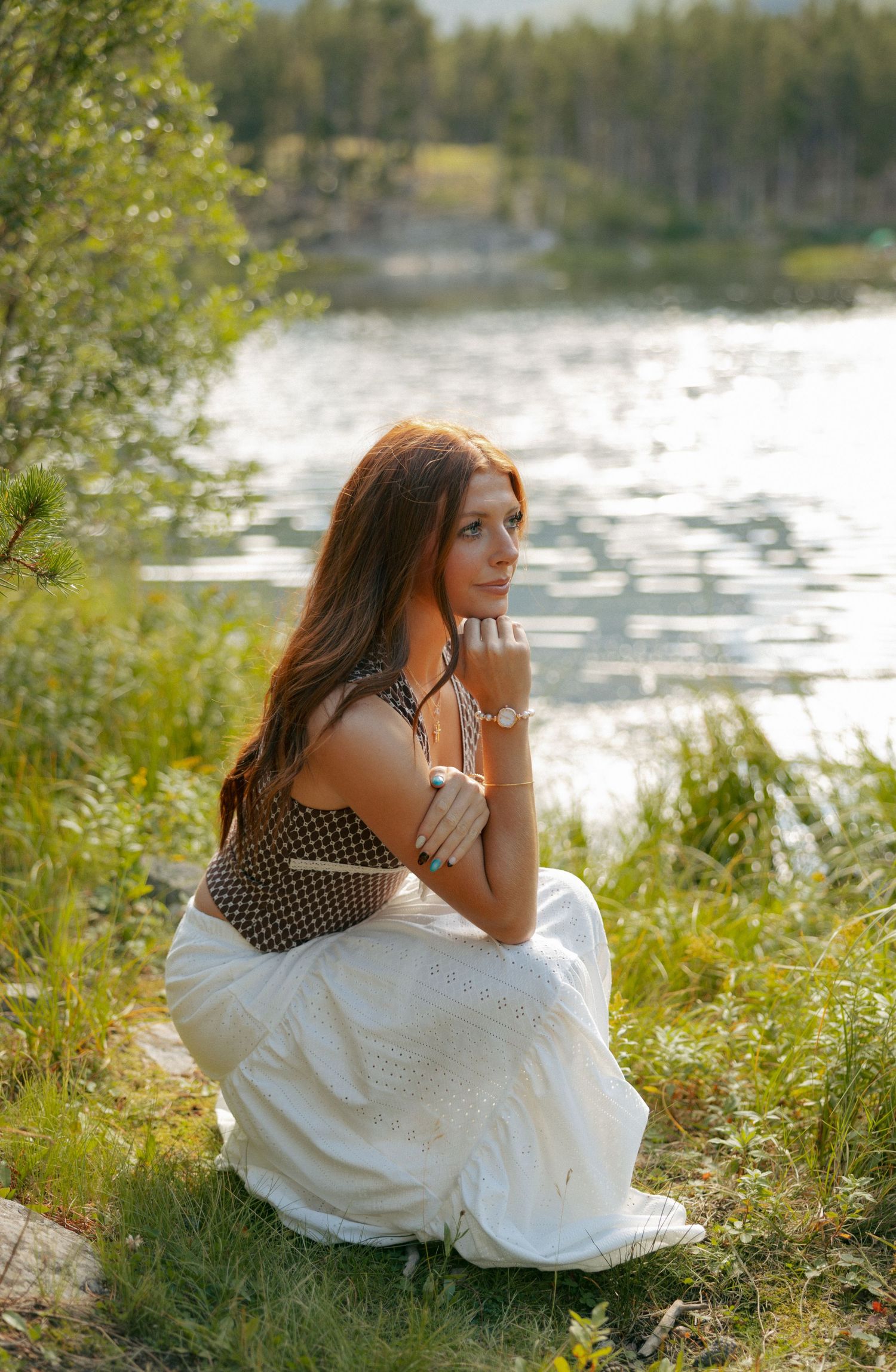 Young woman in white skirt and dark top sits by a serene lake surrounded by lush greenery in golden sunlight.