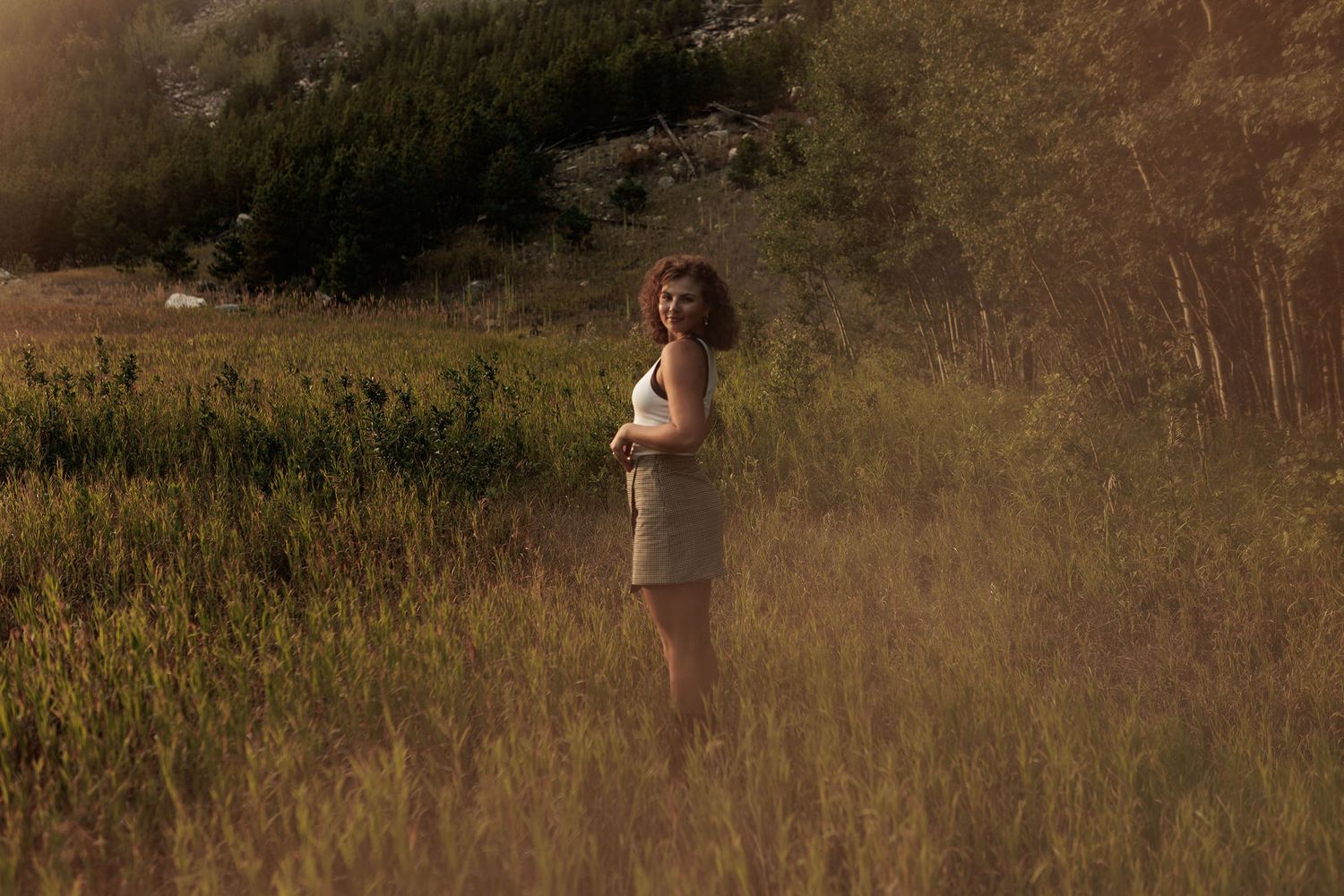 Young woman in white dress standing alone in a sunlit golden meadow surrounded by tall green grass and trees.
