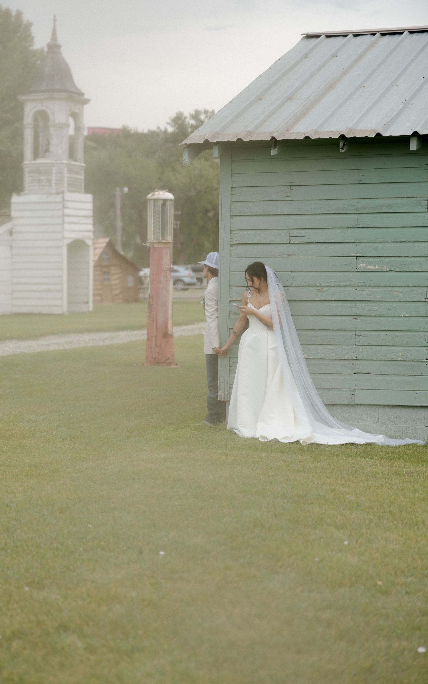 Bride in white gown and veil stands by green shed as groom approaches during outdoor first look.