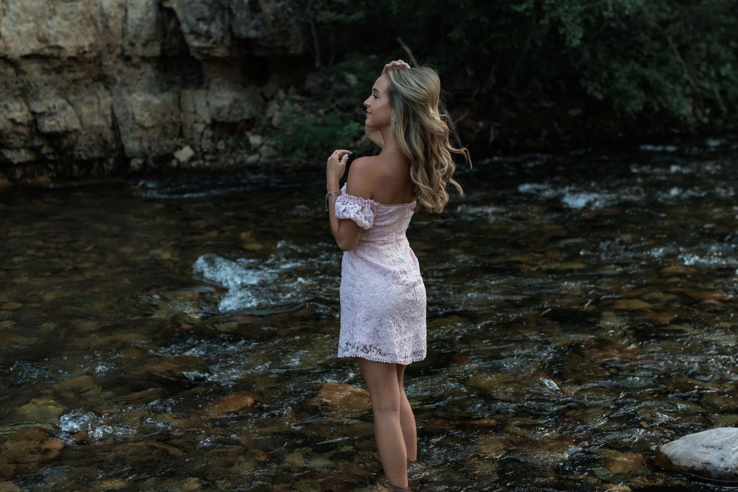 Blonde woman in a pink off-shoulder dress standing in a rocky creek with lush greenery.