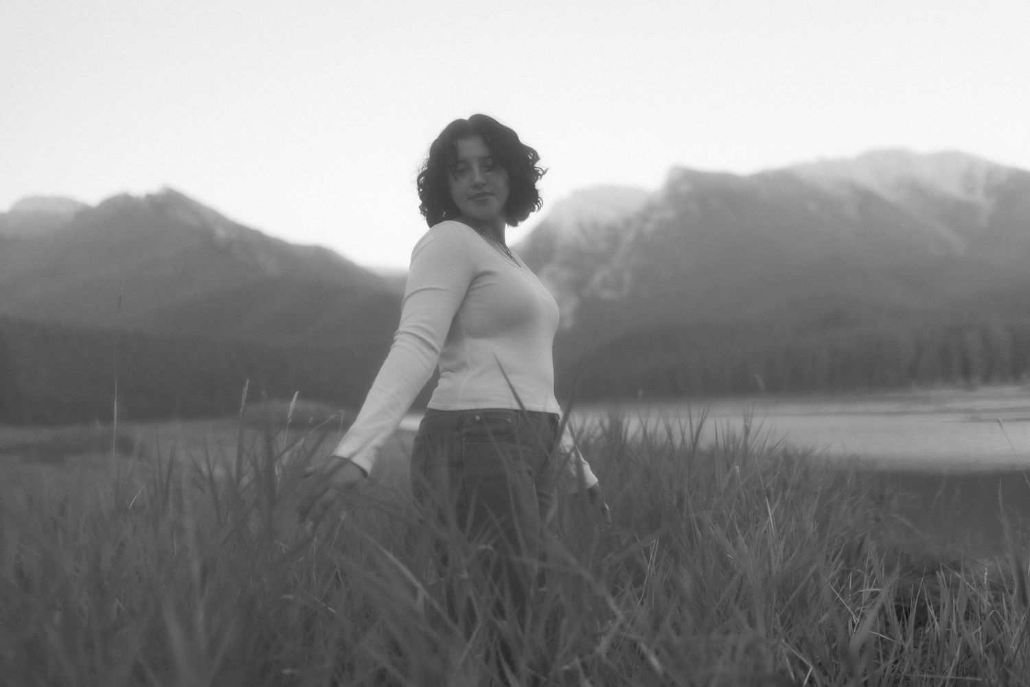 Black and white photo of a curly-haired woman standing in tall grass with mountains and lake in the background.