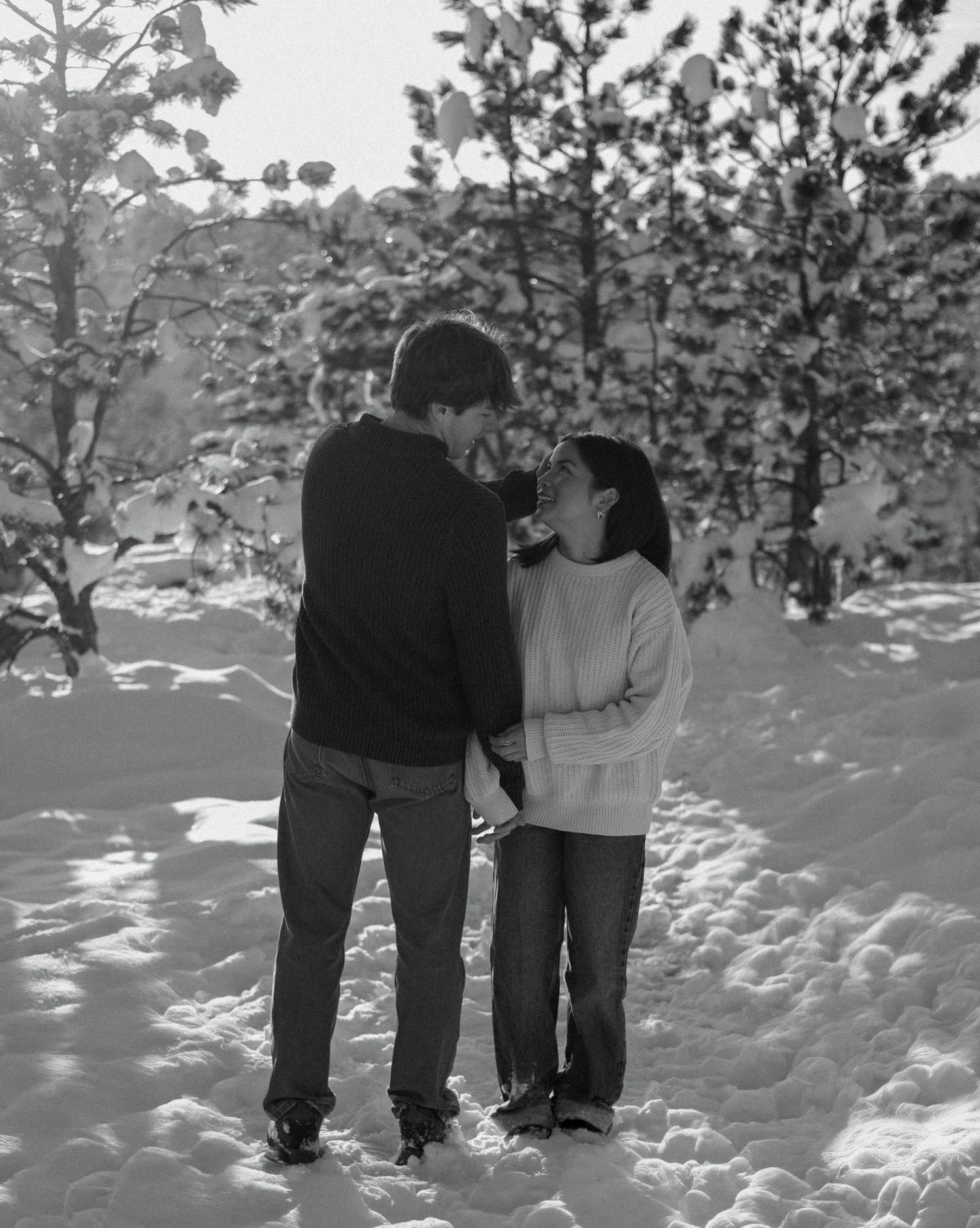 Black and white photo of a couple standing close together in a snowy forest, nearly kissing.