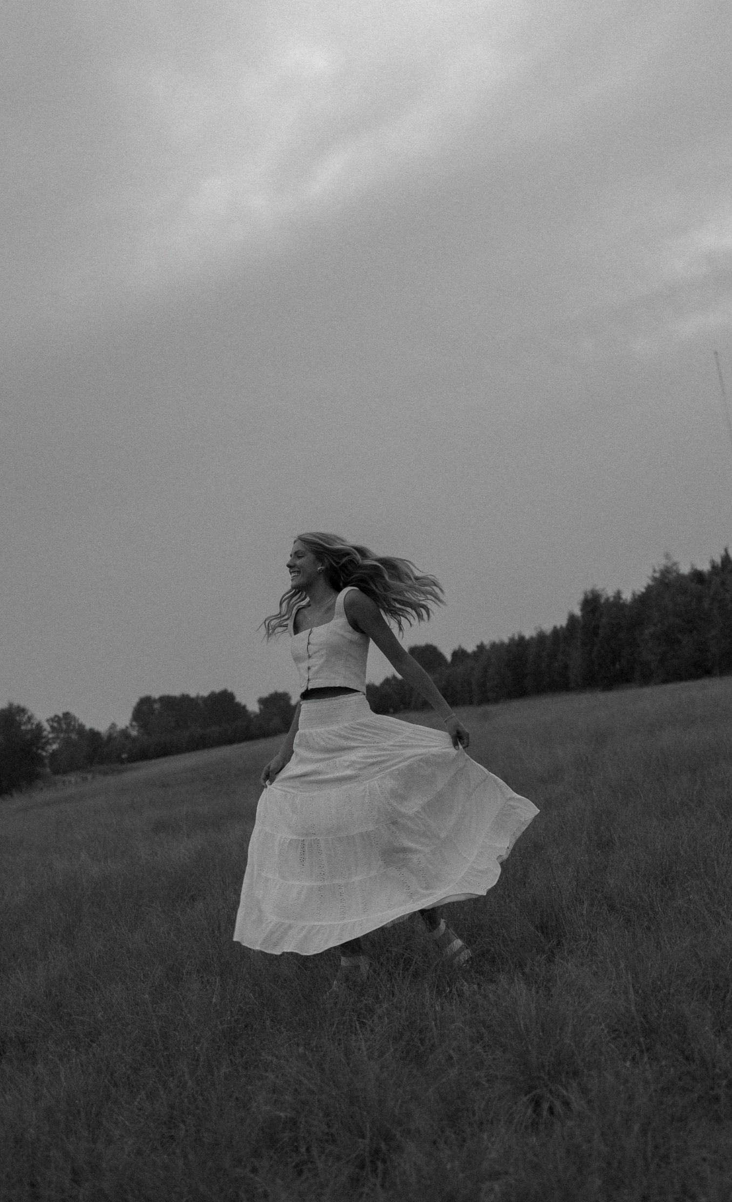 Black and white photo of a woman in a flowing white dress running through an open field under a cloudy sky.