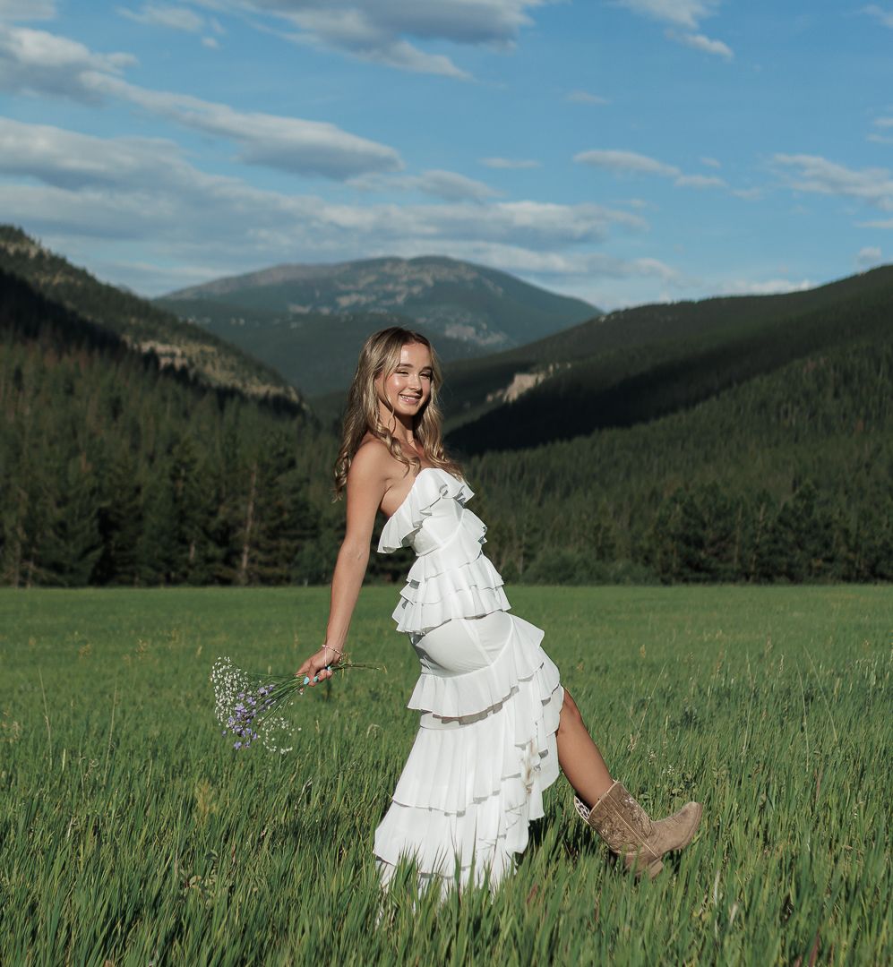 Young woman in white tiered ruffle dress walking through green mountain meadow.