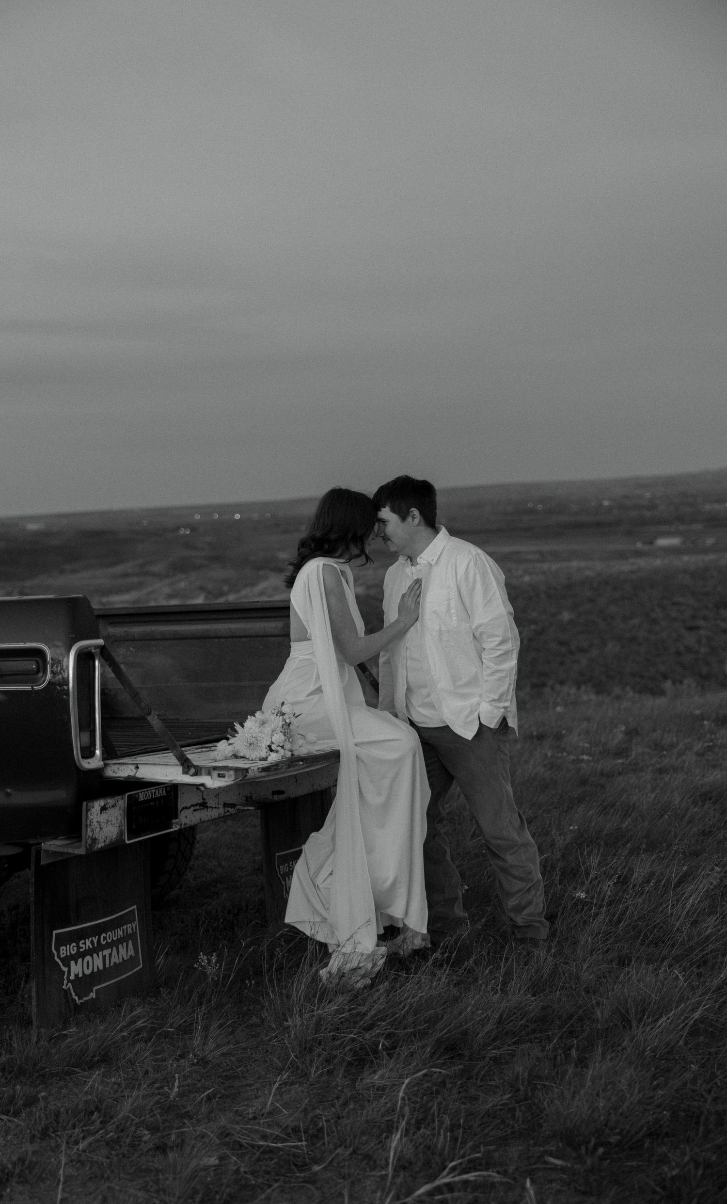 Black and white photo of a bride and groom sharing a romantic moment by a Montana truck in an open field.