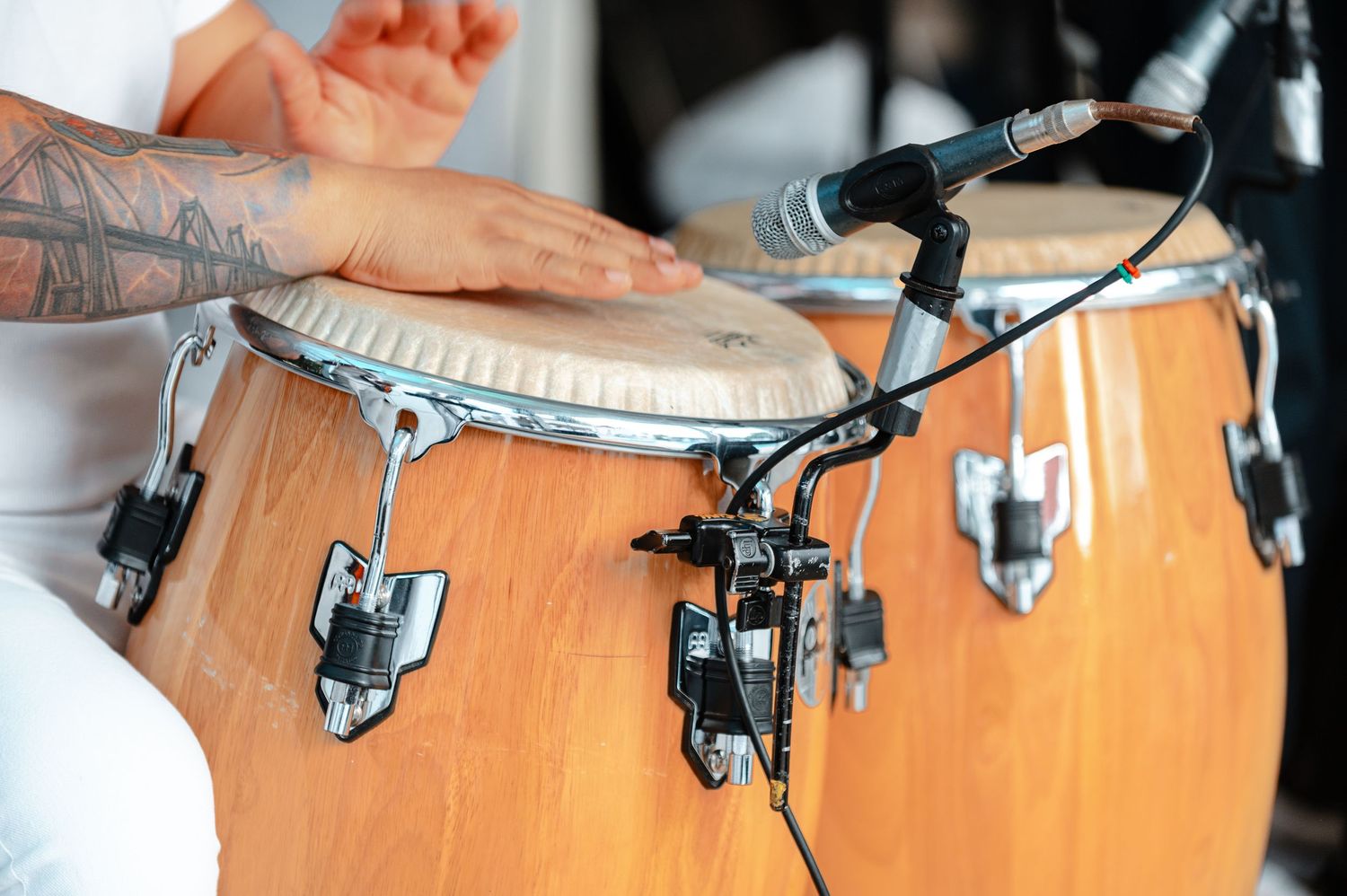 Tattooed hands playing wooden conga drums with microphone at live music event.