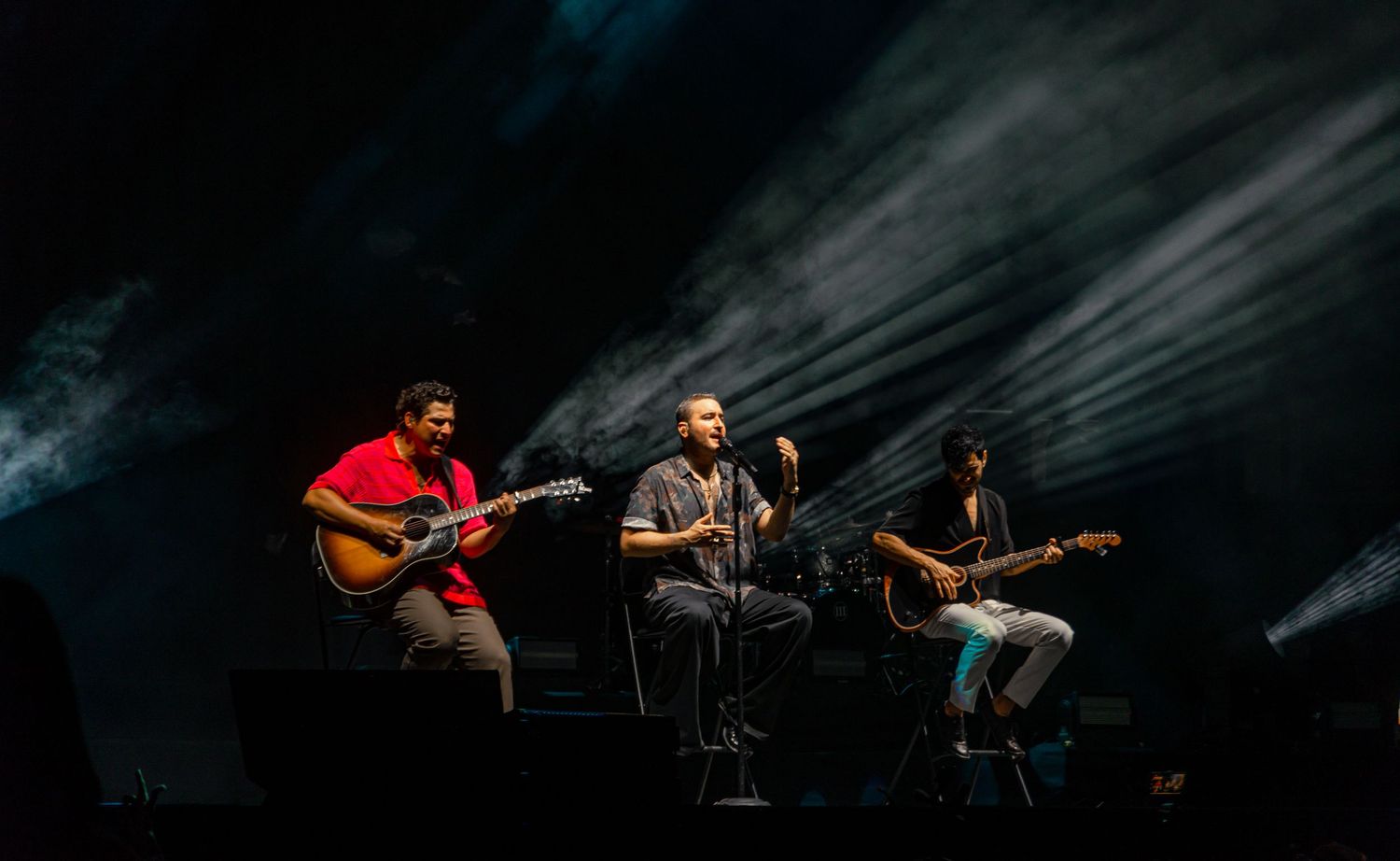 Three musicians performing on a dark stage, two with acoustic guitars and one singing.