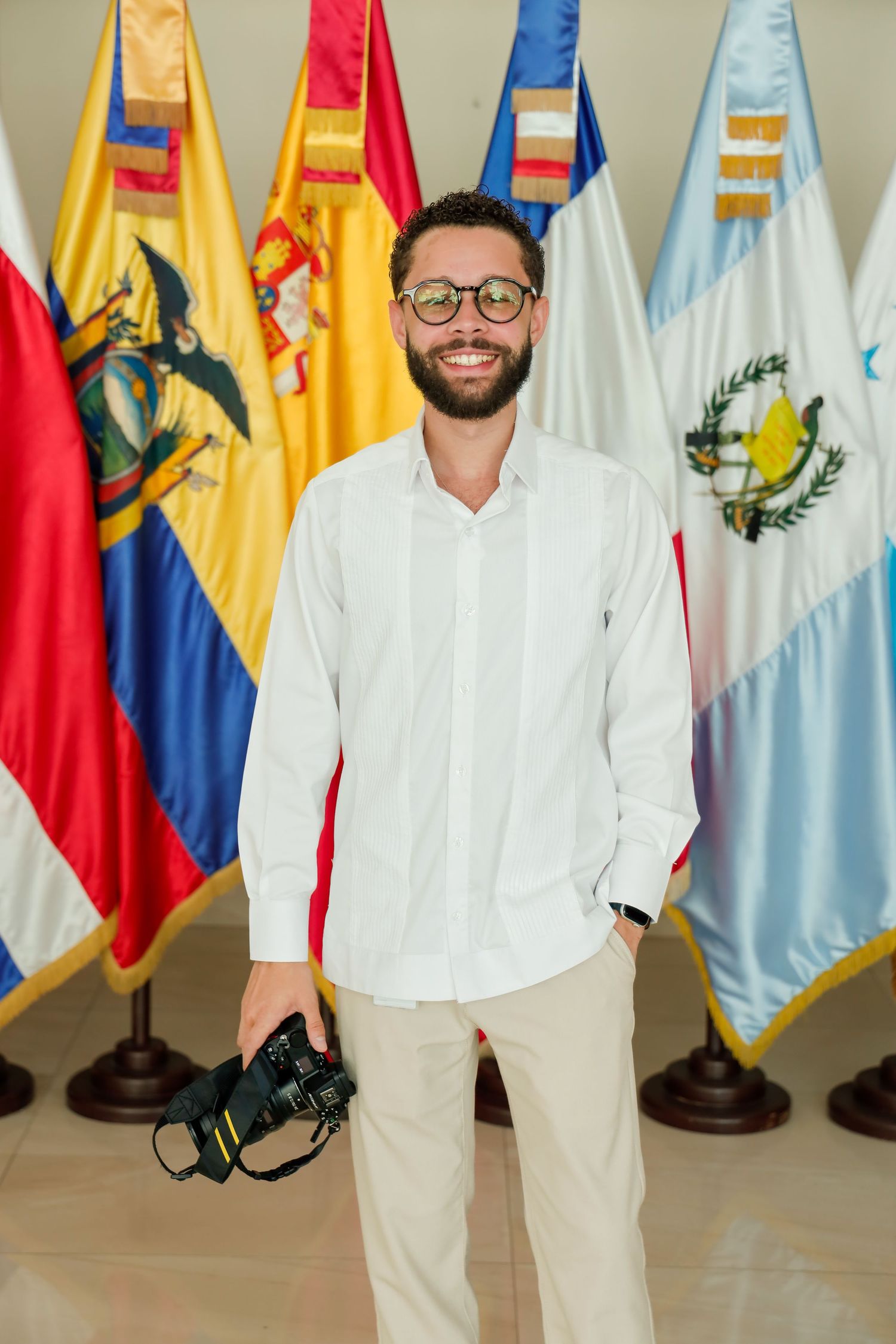 Smiling bearded man holding camera stands before a row of Latin American country flags.
