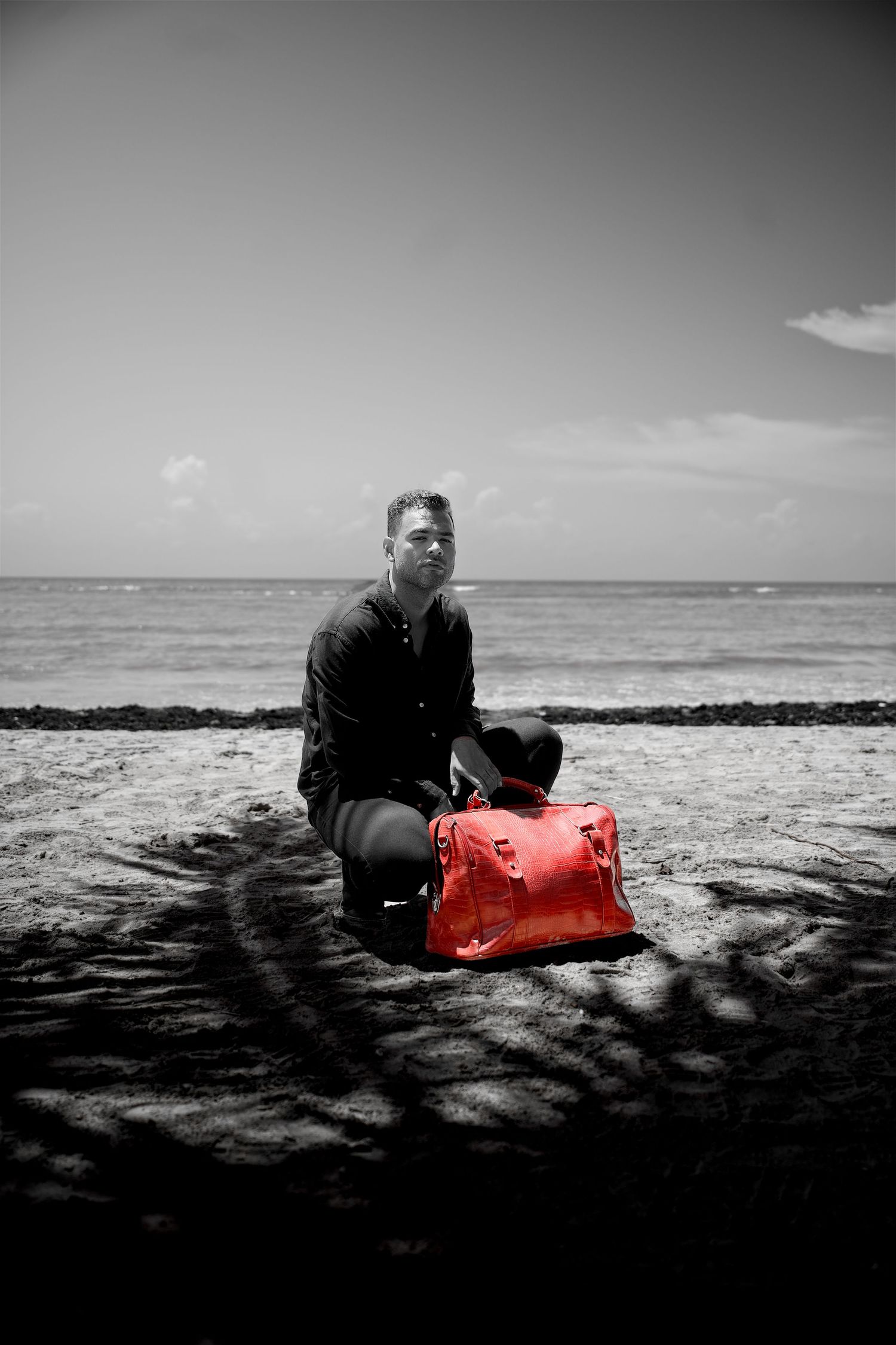 Black and white photo of man kneeling on beach with vibrant red Nike duffel bag by ocean.