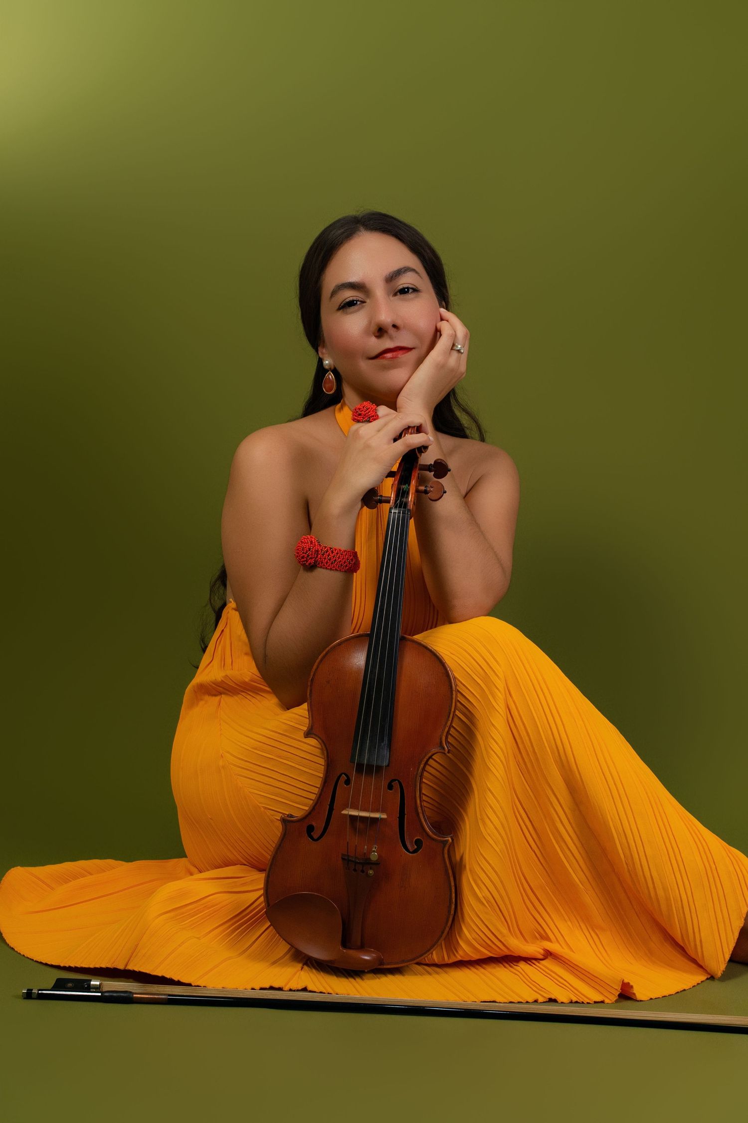 A smiling woman in a yellow dress sitting with a violin against an olive green studio background.