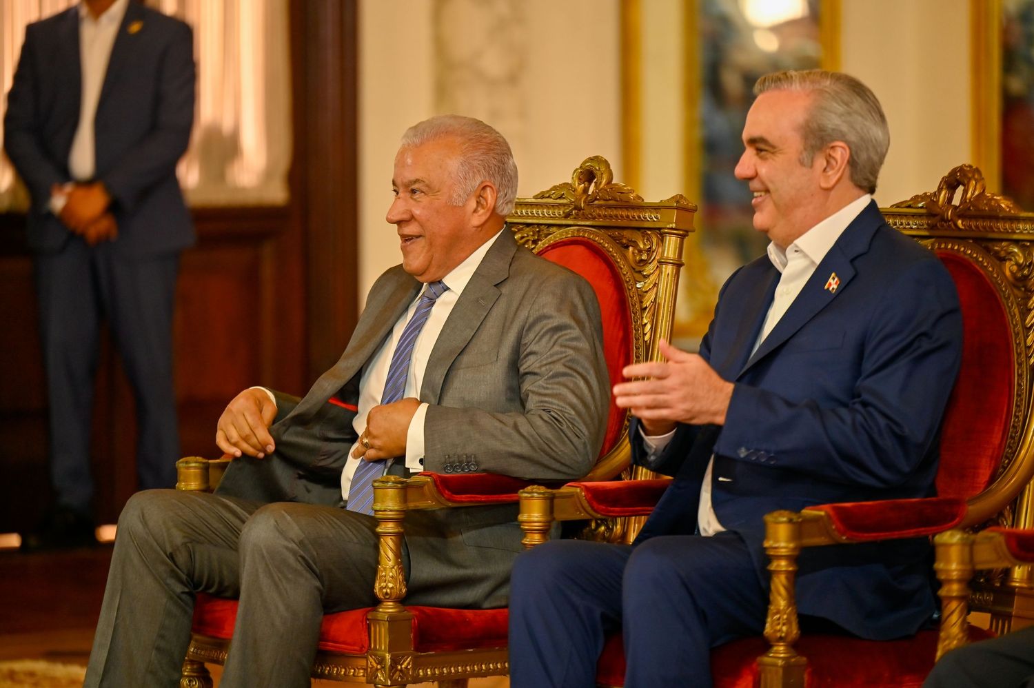 Two men in suits sitting on ornate gold chairs smiling and conversing at formal event.
