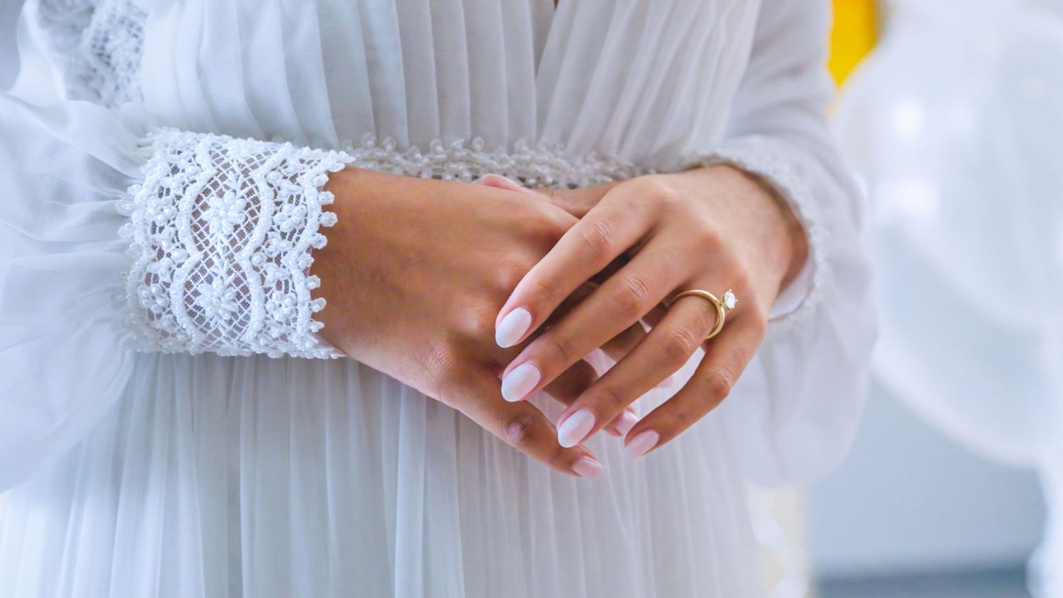 Close-up of bride's hands with gold wedding ring and white lace dress sleeve.