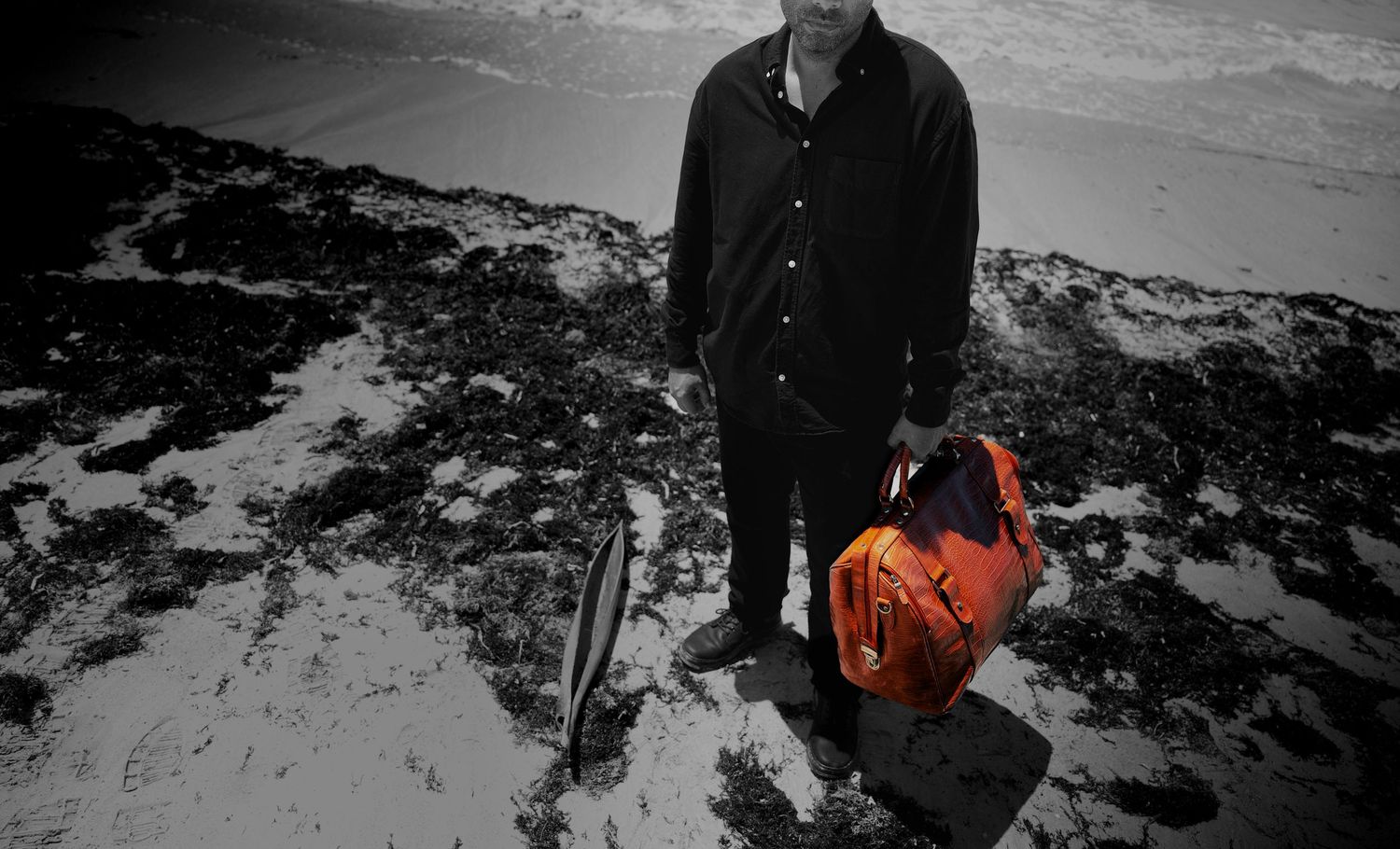 A man standing on a rocky beach holding a bright orange bag in a dramatic black and white landscape photo.