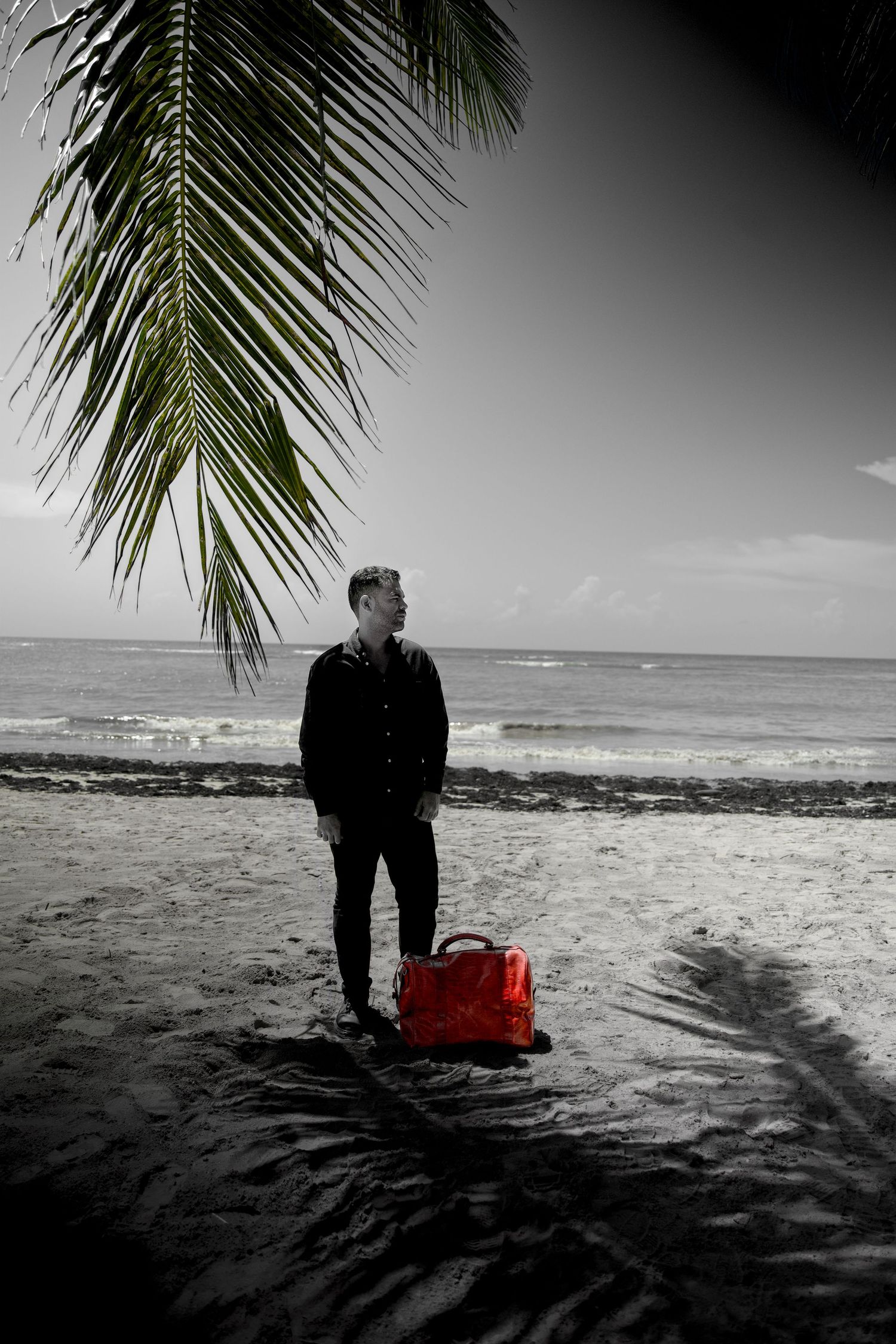 Man standing on black and white beach with vibrant red bag under palm tree by ocean.