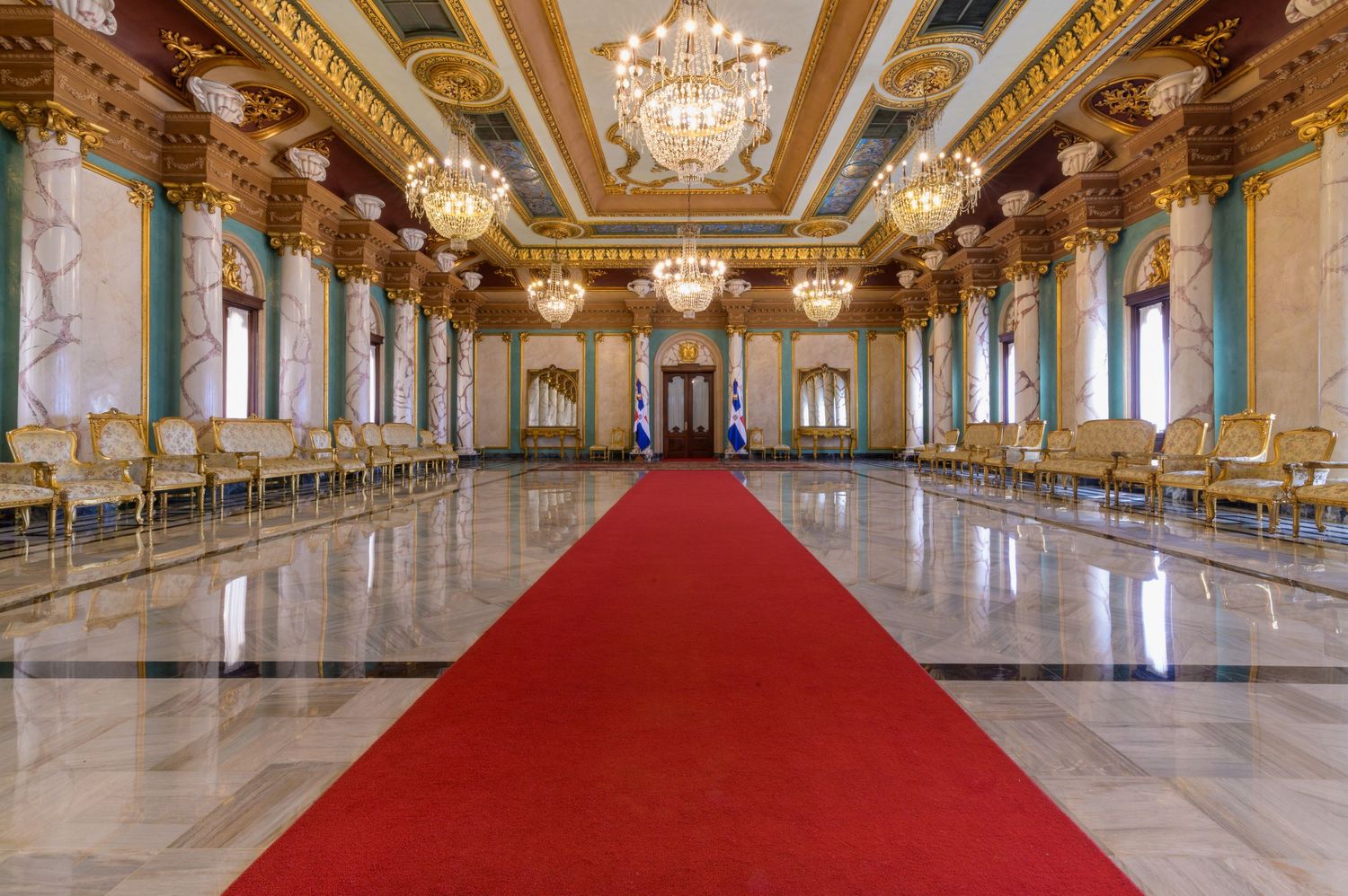 Elegant ballroom with red carpet runner, crystal chandeliers, gold accents, and rows of chairs along marble walls.