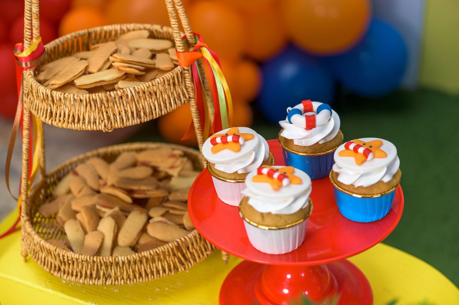 Colorful birthday party dessert table with decorated cupcakes on red stand and cookies in wicker basket.