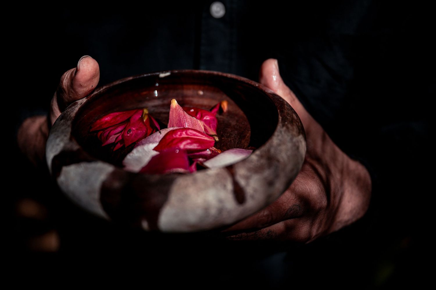 Hands holding a rustic bowl with red flower petals and a small candle flame in dark atmospheric lighting.