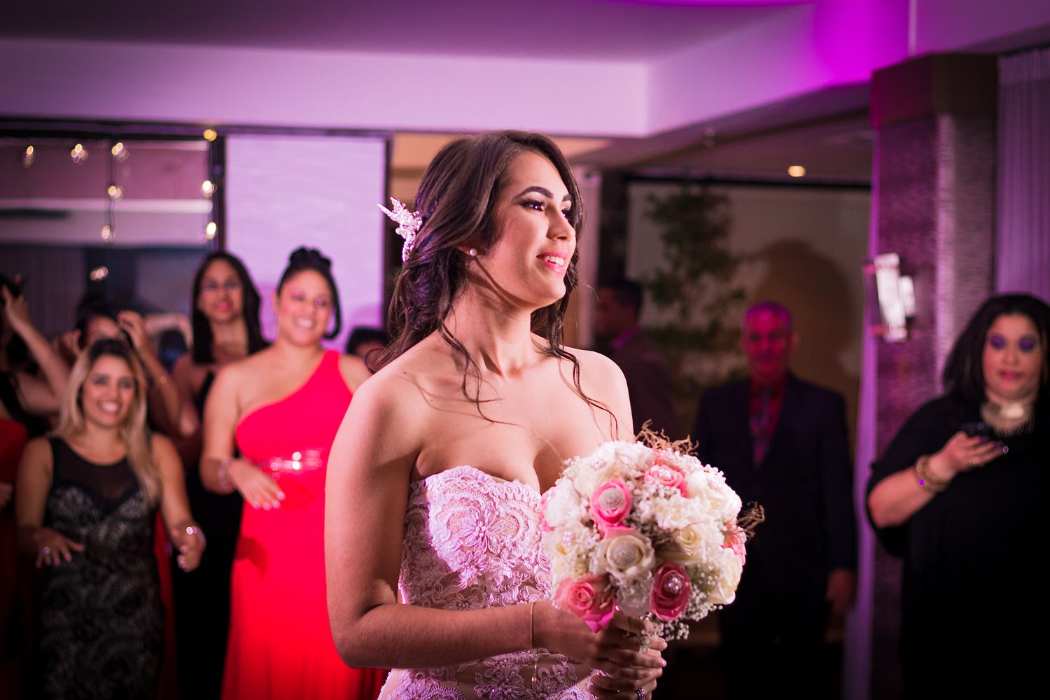 Bride in strapless lace gown preparing to toss bouquet to excited guests at reception.