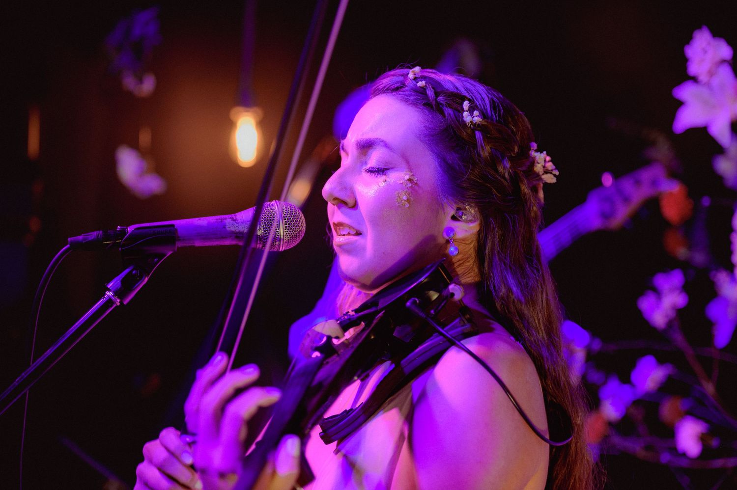 Female vocalist singing into a microphone surrounded by flowers under purple stage lighting.