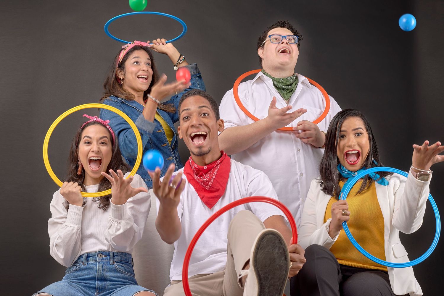 Group of five diverse friends posing playfully with colorful hula hoops on dark studio background.