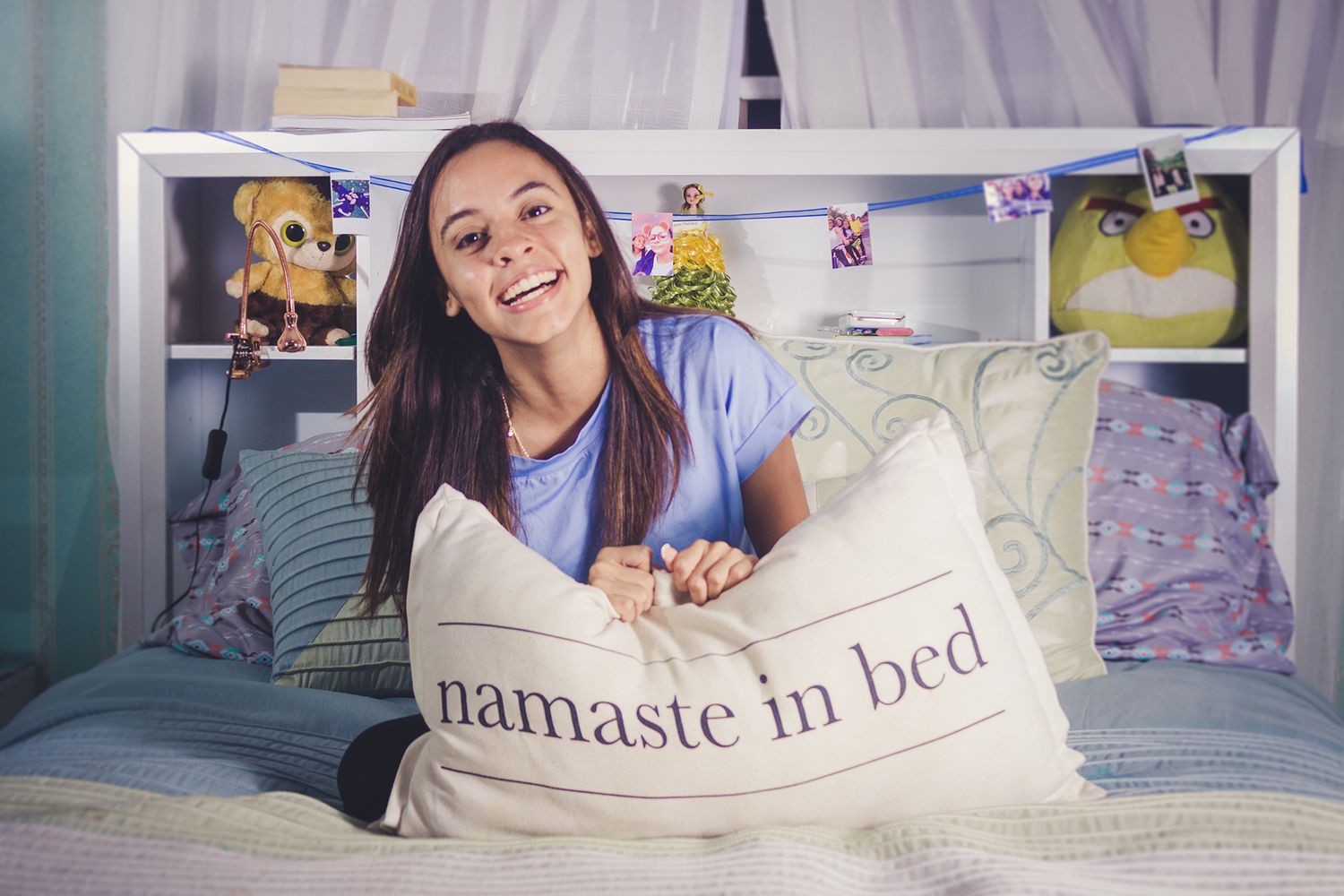 Smiling young woman hugging a namaste in bed pillow while sitting on a cozy decorated bed.