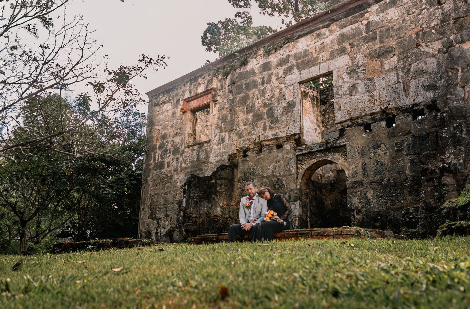 Couple sitting together on grass before historic stone ruins with bouquet.