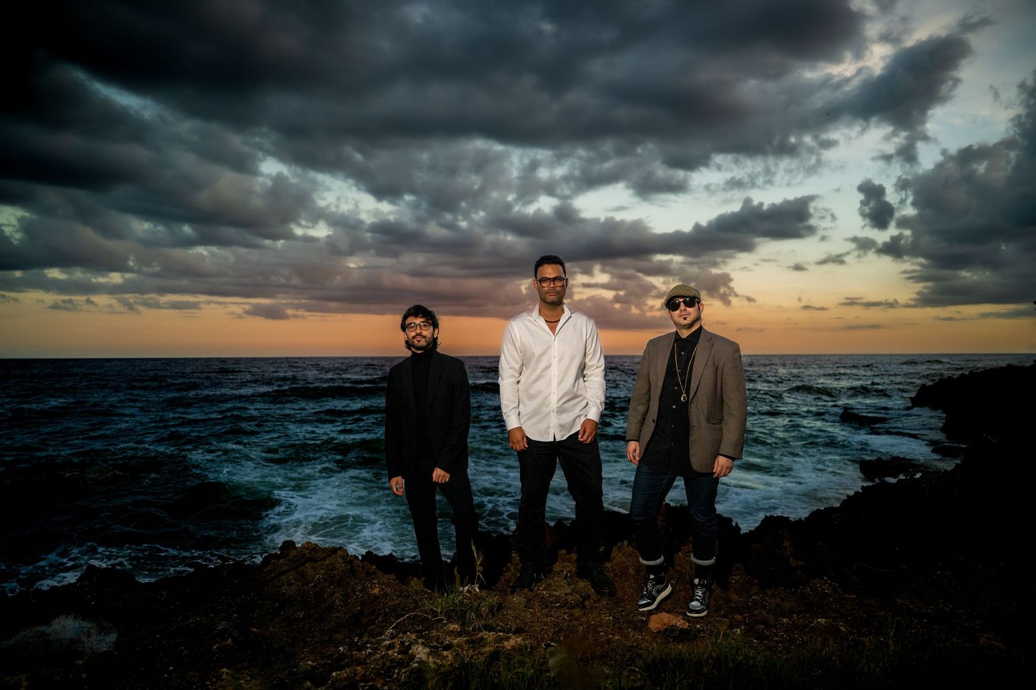 Three men standing on a rocky shoreline during a vibrant sunset with dramatic storm clouds overhead.