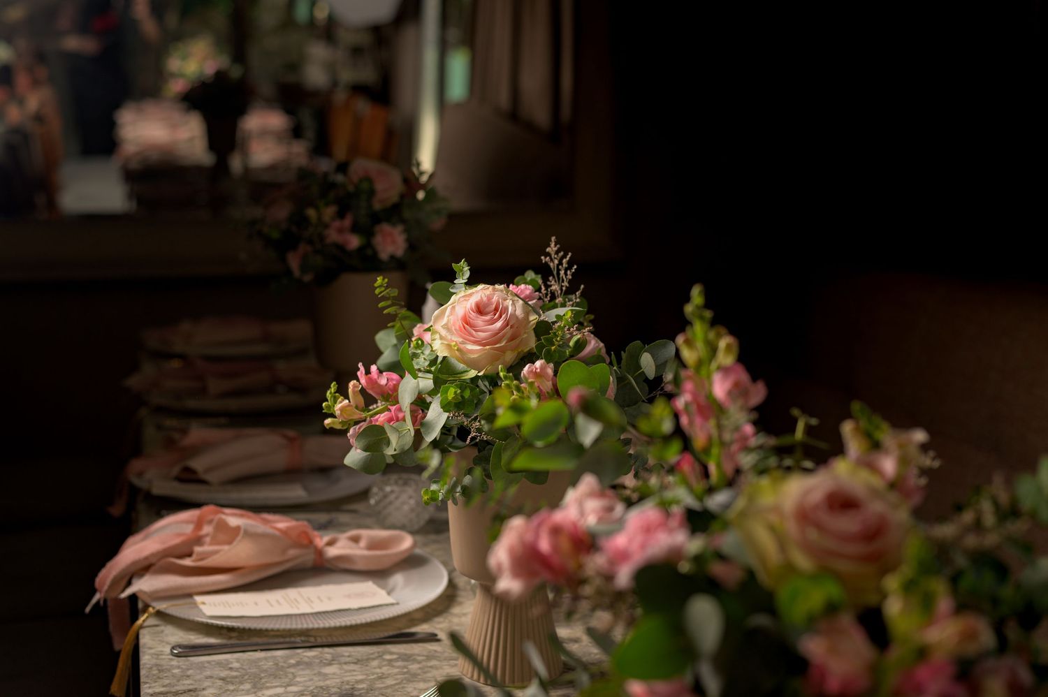 Elegant wedding table setting with pink roses and greenery floral arrangements in vintage vases on a linen tablecloth.