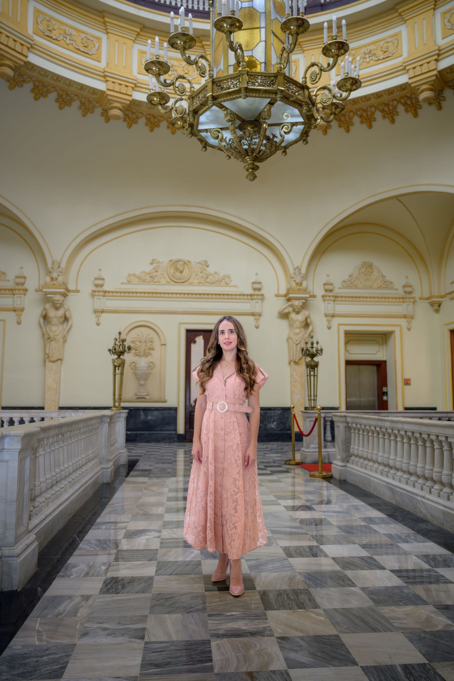 Elegant woman in pink lace dress standing in ornate classical building with chandelier.