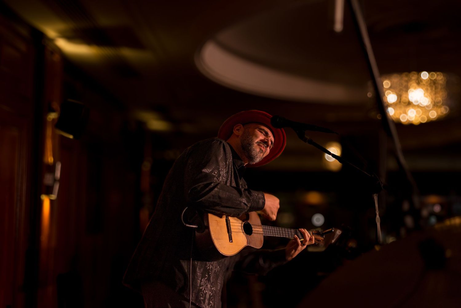 Musician playing acoustic guitar on a dimly lit stage wearing a red hat.
