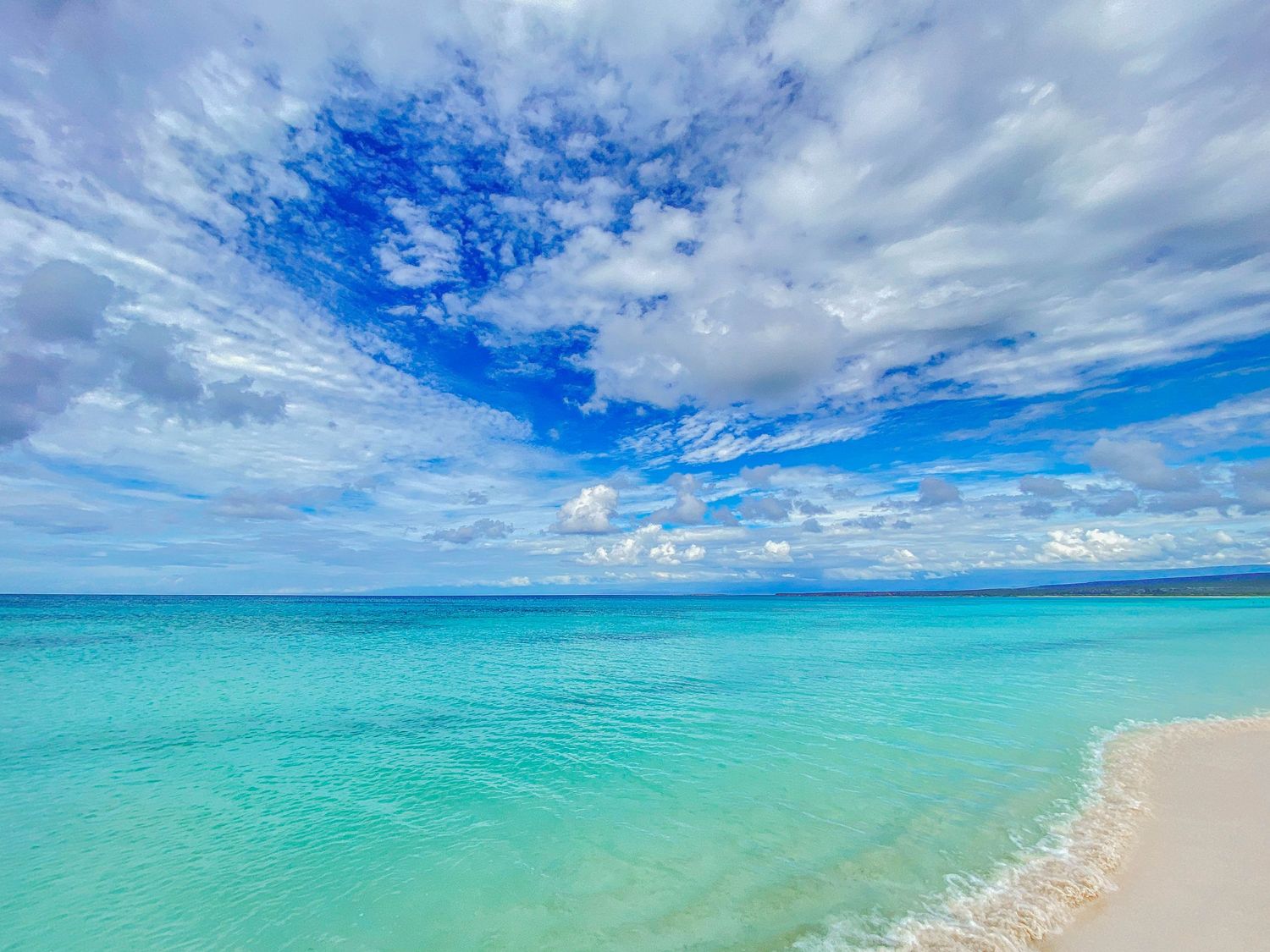 Crystal clear turquoise ocean water meets a white sandy beach under a vivid blue sky with scattered white clouds.