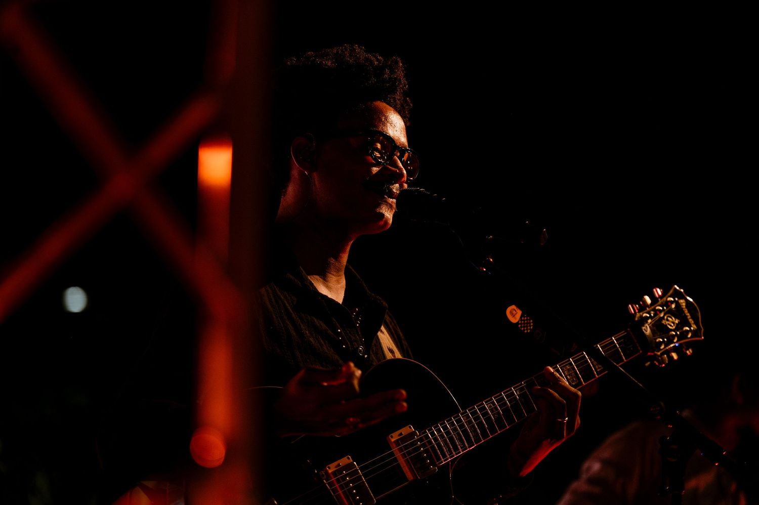 A musician playing an electric guitar on a dimly lit stage bathed in warm red light.