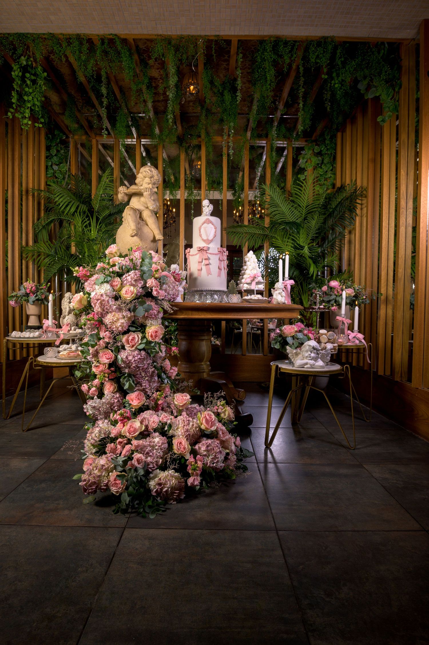 Stunning pink floral dessert table with tiered cake and angel statue under greenery.