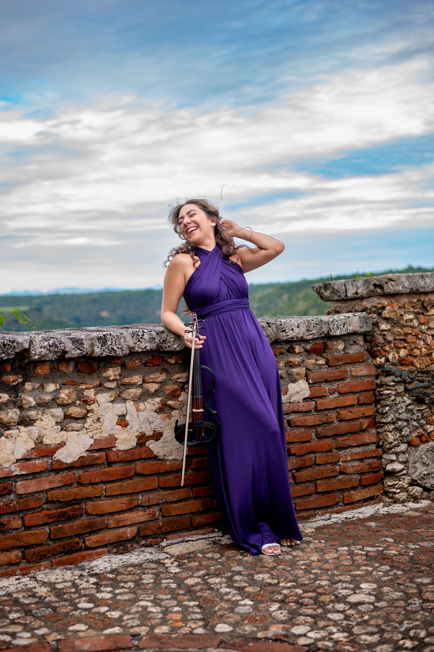 Woman in purple gown holding violin bow on rustic brick terrace with scenic sky backdrop.