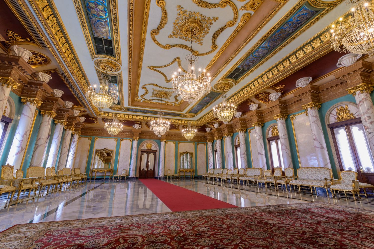 Opulent baroque ballroom featuring elaborate painted ceiling, crystal chandeliers, red carpet, and gilded white chairs.