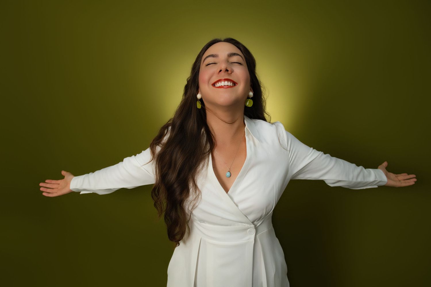 Happy woman in white wrap dress with arms outstretched on olive background.
