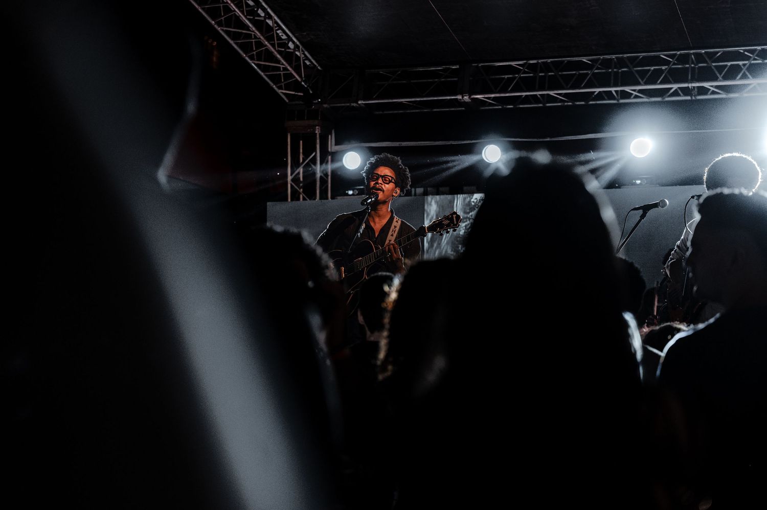 Guitar player performing on an outdoor stage at night with bright spotlights behind them.