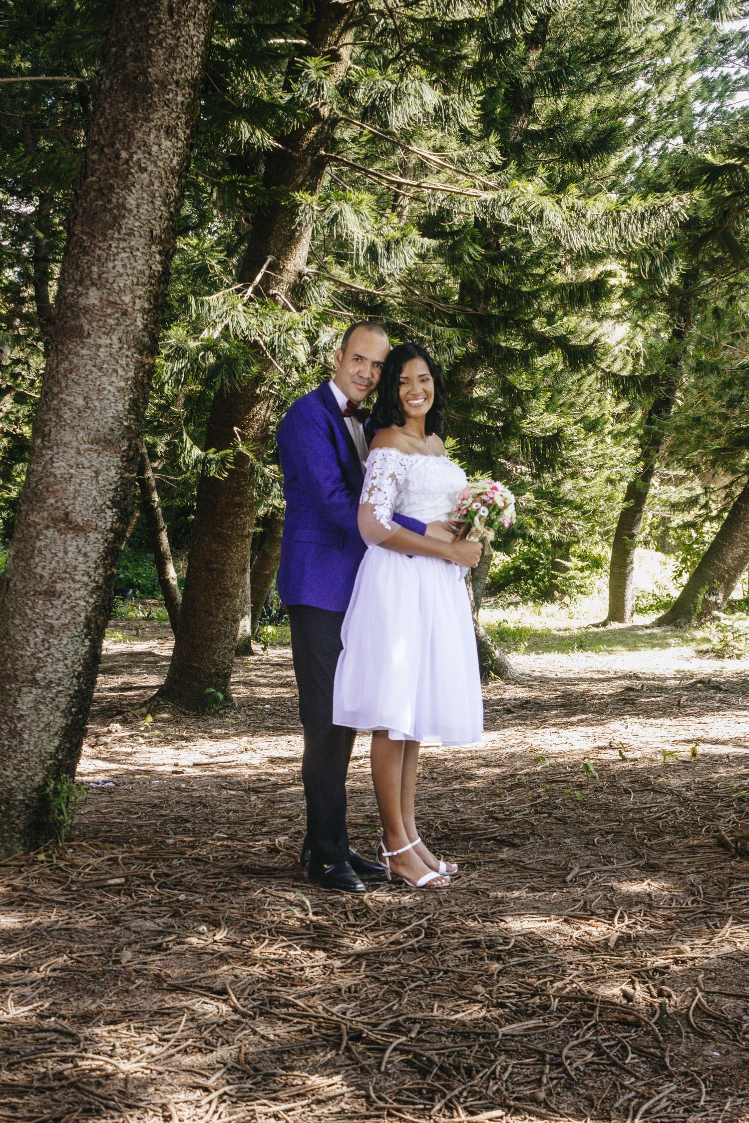 Bride in white dress and groom in purple tuxedo posing together in a sunlit forest setting.