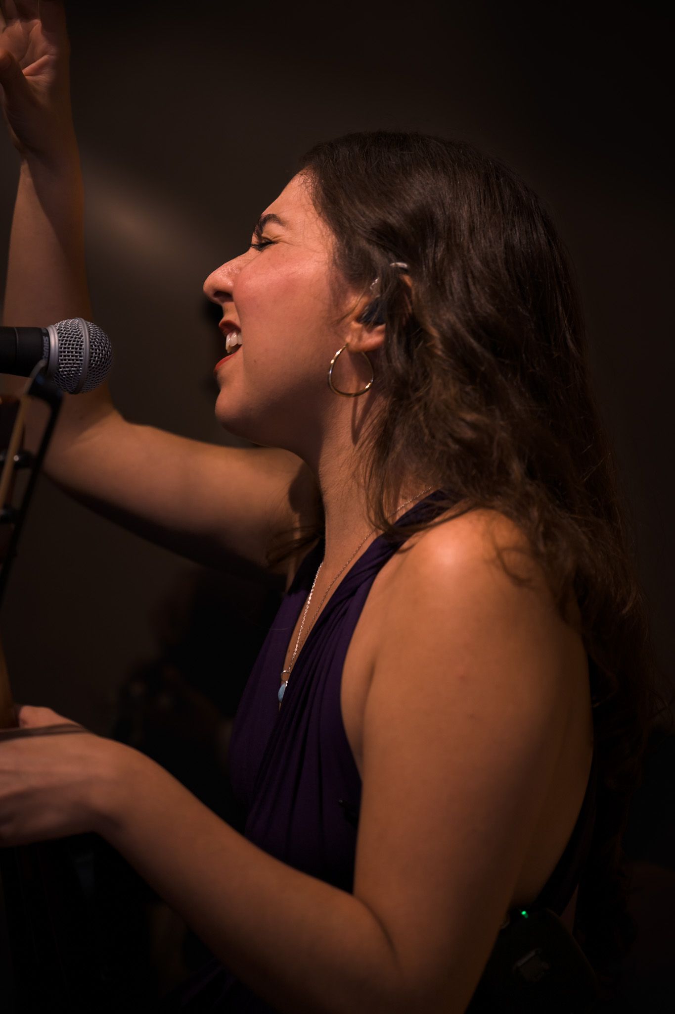 Young woman singing passionately into microphone on stage wearing purple top in dim lighting.