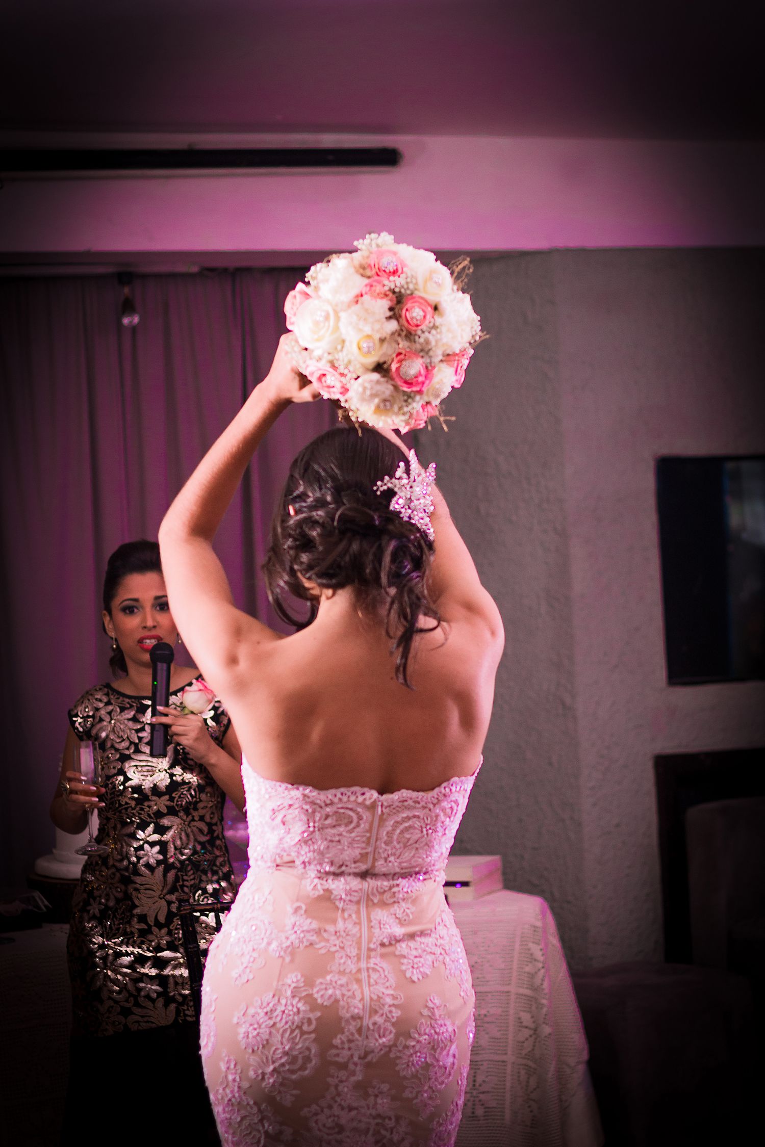 Bride in lace wedding gown raising floral bouquet overhead preparing for bouquet toss at reception.