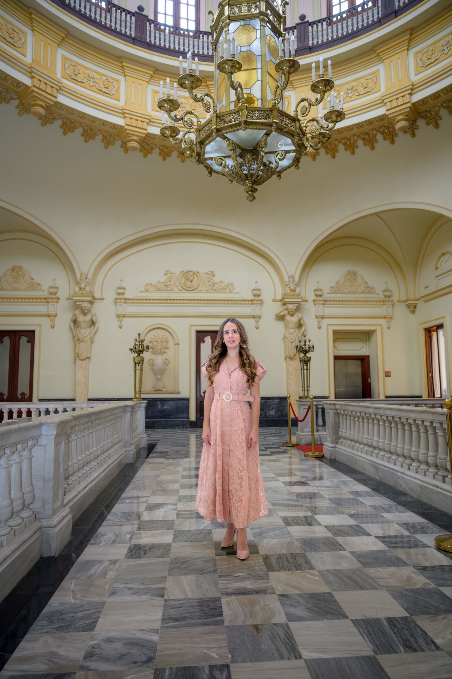 Woman in pink lace midi dress standing in elegant neoclassical building with ornate chandelier.