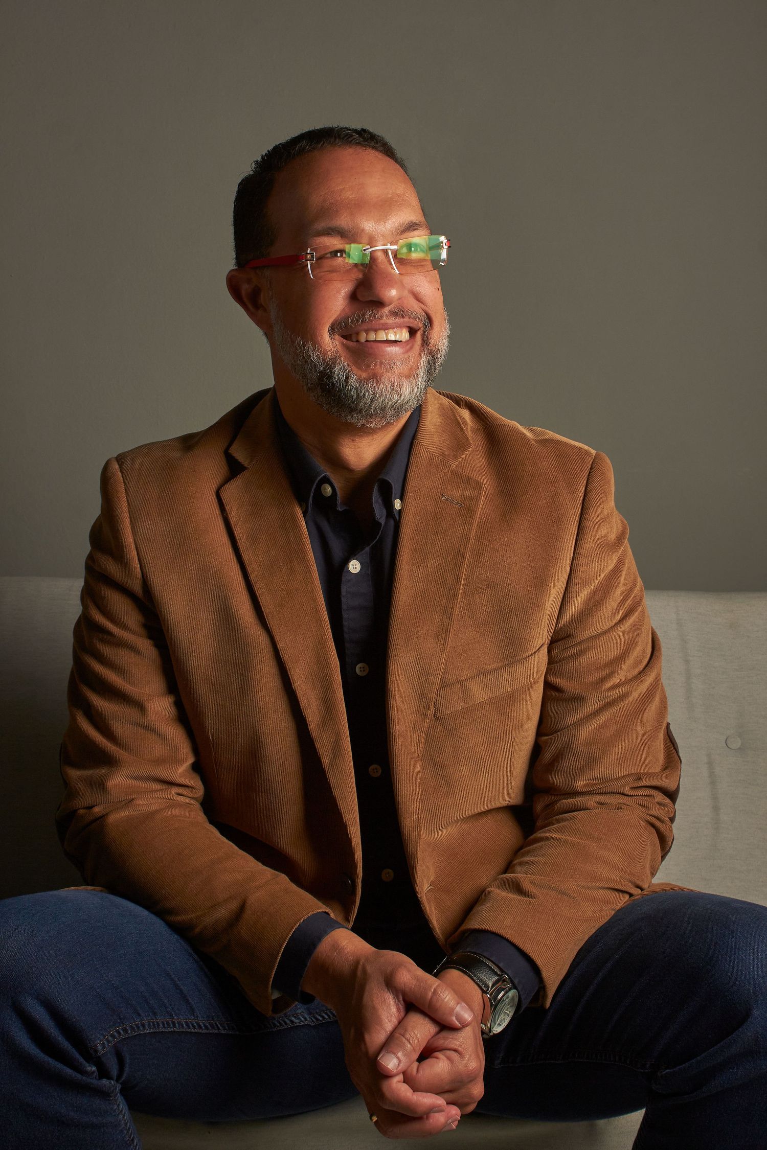 Smiling man in brown blazer and colorful glasses sitting on a couch with hands clasped.